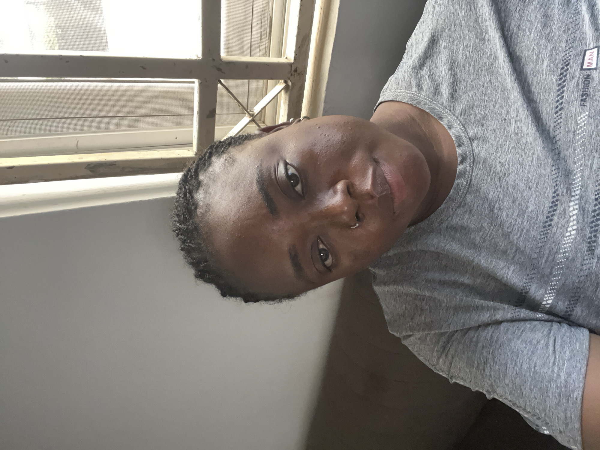 A young Black person with dark braided hair and a nose ring is captured in a portrait, lying on their side. They are wearing a gray t-shirt with a subtle pattern and the words "FASHION MAN" printed vertically on the right side. The person is looking directly at the camera with a relaxed expression.
The background suggests an indoor setting with natural light streaming in from a window. The window has a white metal frame, and behind it, a white screen is visible, indicating it's likely a warm climate. The overall lighting is bright, suggesting it might be daytime. The wall behind the person appears to be painted a neutral gray. The composition is close-up, focusing on the individual's face and upper body.
The image appears to be a selfie, taken from a low angle, creating an intimate feel. The person's skin is well-lit, with a slight sheen, possibly due to sweat or a dewy complexion. The focus is sharp on the face, highlighting the person's features and jewelry. The image conveys a sense of casualness and self-expression.