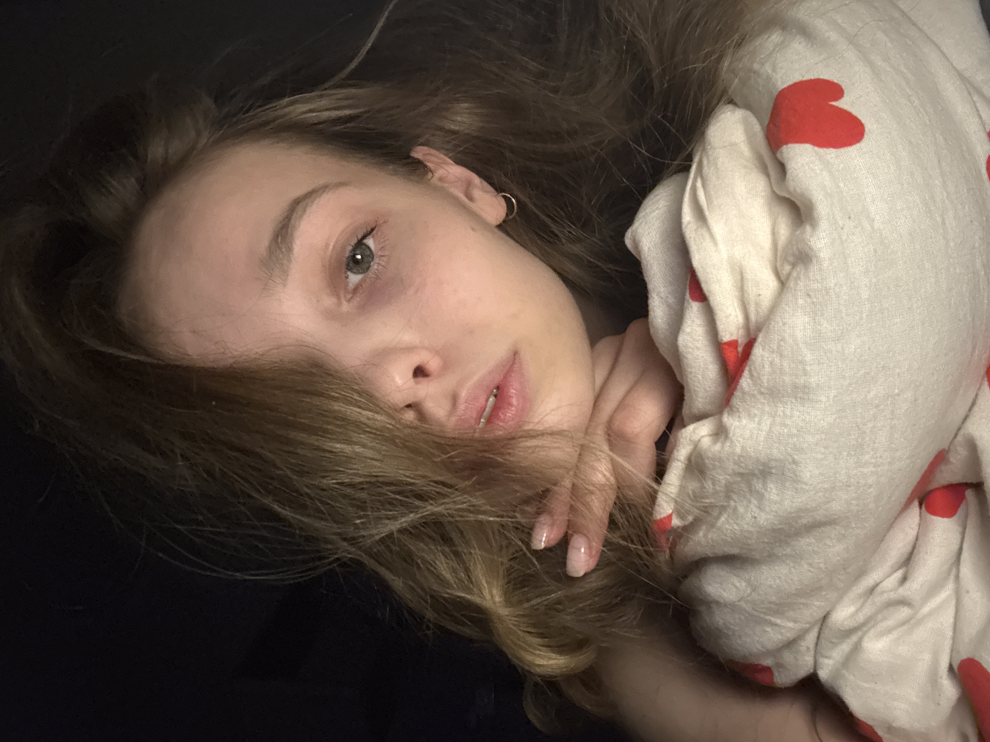 A fair-skinned young woman with light brown hair is lying on a dark surface, possibly a bed. Her head is tilted towards the viewer, and her hair partially obscures her face. She has light eyes and pink lips. She is wearing a small hoop earring in her left ear. Her hand is near her face, with her fingers visible. She is wrapped in a light-colored blanket or sheet with red heart-shaped patterns. The background is dark and indistinct. There is no information available about the location of the image in the city of Chaikovsky, Russia.