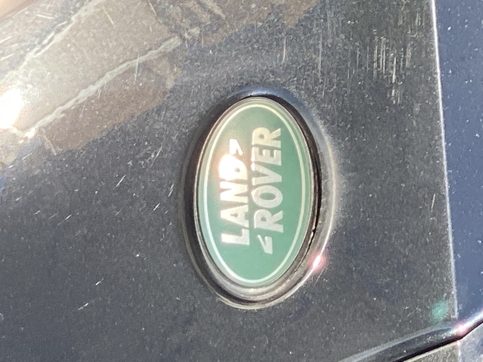 The image shows a close-up of the Land Rover logo affixed to the exterior of a vehicle. The oval-shaped logo features the words "LAND ROVER" in a light color against a green background. The vehicle's surface around the logo appears to be dark and slightly textured with signs of wear or dirt. The photograph was not taken in Budva, Montenegro.