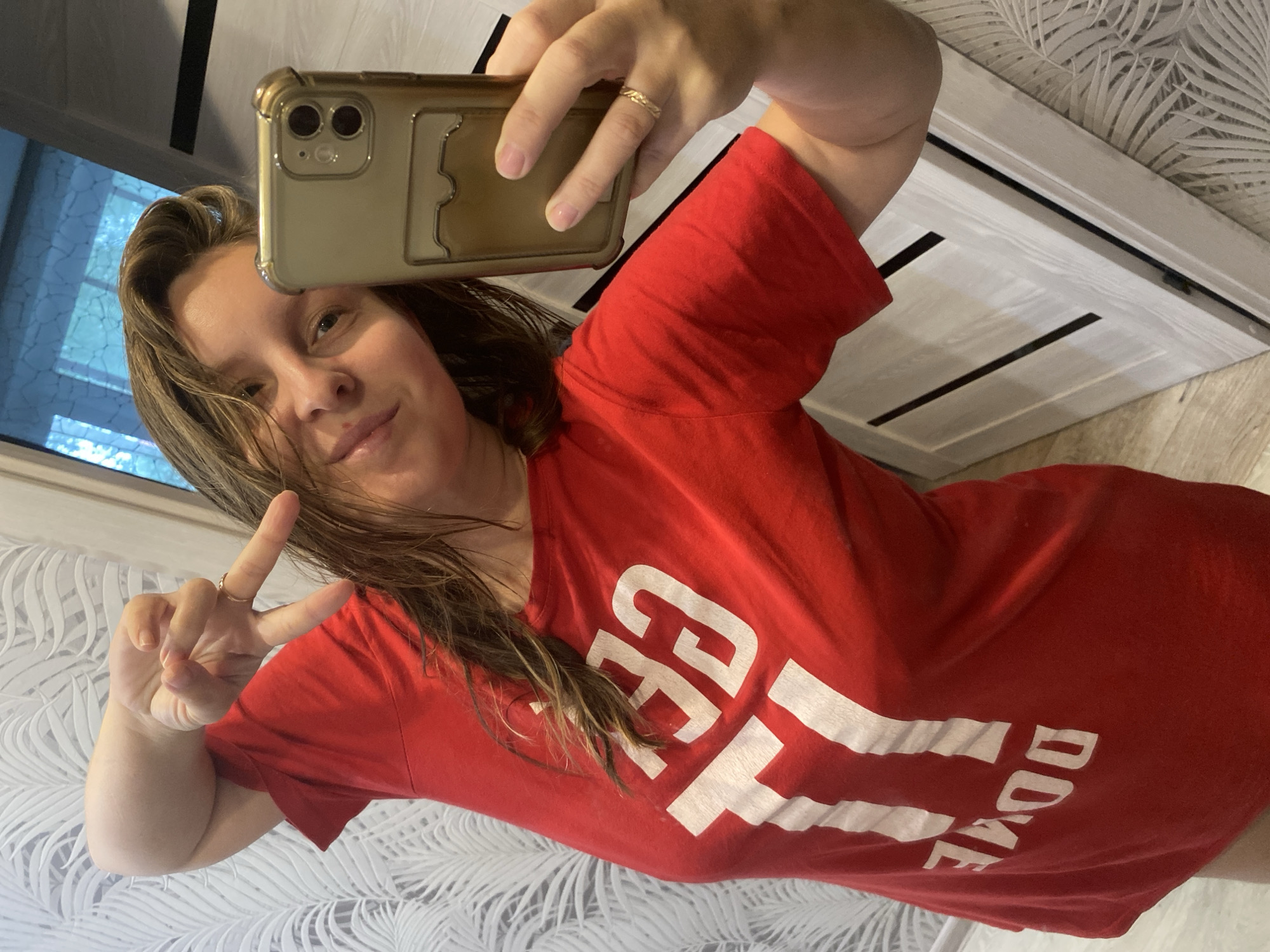 A woman with medium-length brown hair, wearing a red t-shirt with white lettering that reads "GET IT DONE," takes a selfie in a room. She is holding a gold-colored phone with a clear case in her right hand, and her left hand is raised in a peace sign gesture. The background shows a patterned wallpaper and a closet door with a diamond-shaped design. The lighting suggests an indoor setting, possibly a home. The image does not contain any information about the specific location in Noviy, Russia.