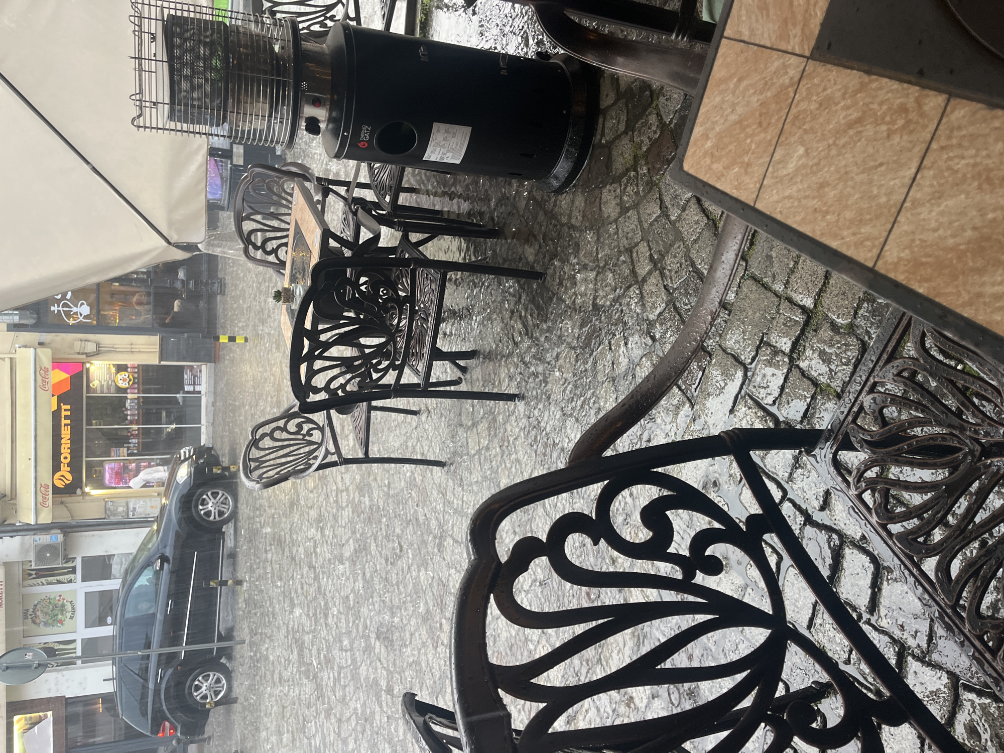 The image shows an outdoor seating area of a cafe or restaurant in the city of Plovdiv, Bulgaria during heavy rain. The cobblestone street is wet. The tables and chairs are made of black metal with ornate designs. There is a black patio heater next to one of the tables. The storefronts of businesses, including a "FORNETTI" shop with Coca-Cola signage, are visible across the street. A dark-colored car is parked on the street. There are no people visible under the covered area.