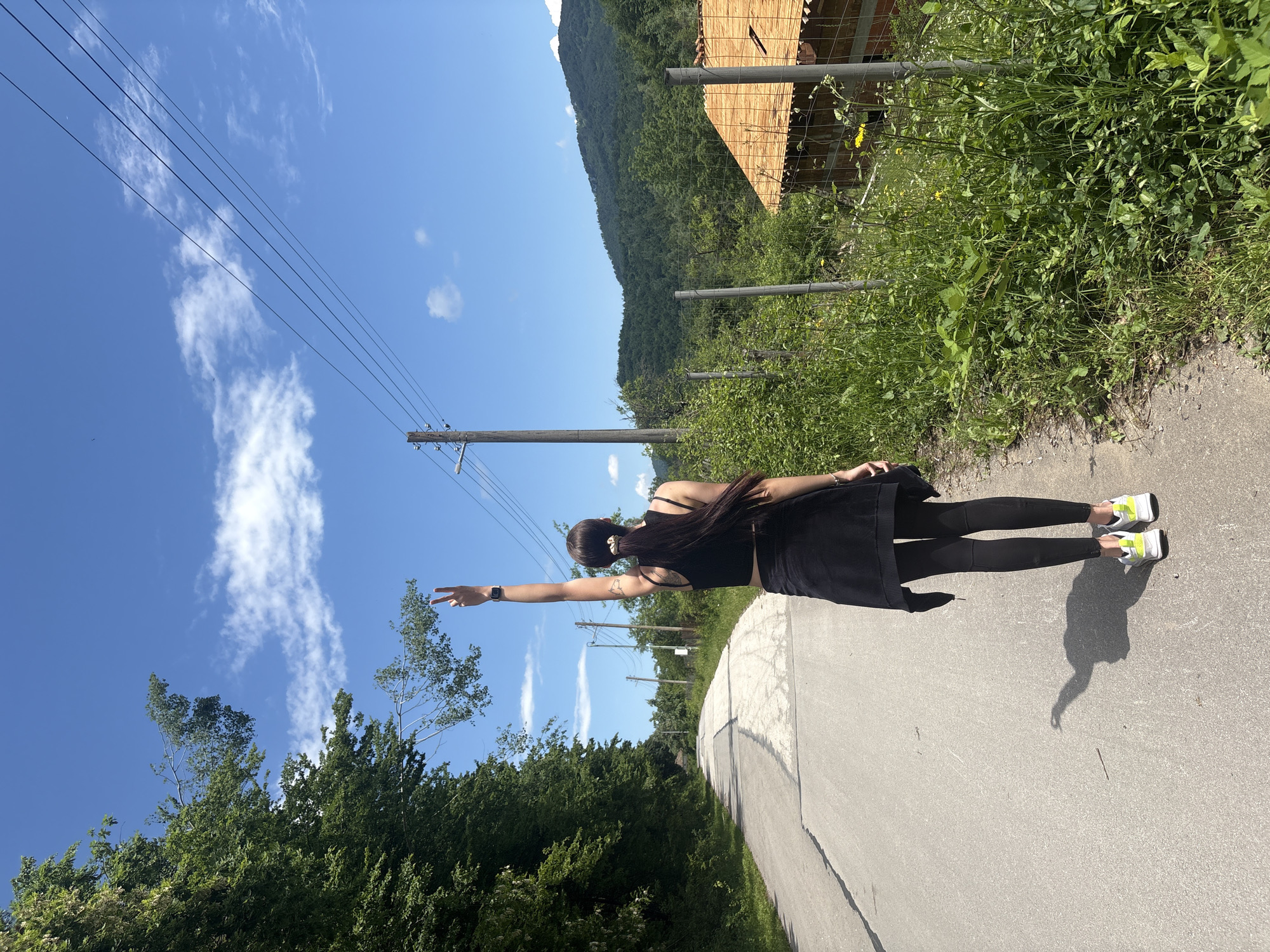 A single individual, viewed from the back, stands on a paved road in a rural setting under a clear blue sky with scattered white clouds. The person, appearing female, wears a black cropped tank top, black leggings, and a black garment tied around her waist. Her long dark hair is styled in a ponytail with a light-colored scrunchie. She wears white sneakers with neon yellow-green accents. Her left arm is raised vertically, with the hand forming a "V" (peace) sign. A smartwatch is visible on her left wrist, and a faint tattoo is on her upper left arm. The sun casts a distinct shadow of the person onto the road behind her, indicating bright daylight.
To the left of the road, a dense line of tall green trees forms a forest edge. The road itself is gray, showing visible cracks and wear, and extends into the distance. To the right, the roadside is densely overgrown with green grass and various plants, including some yellow wildflowers. A wire fence with metal posts runs parallel to the road. Beyond the fence, a partially constructed building with a terracotta-tiled roof is visible. Further in the background, a series of verdant hills or mountains rise. A wooden utility pole with multiple horizontal power lines crosses the upper right portion of the image against the sky.