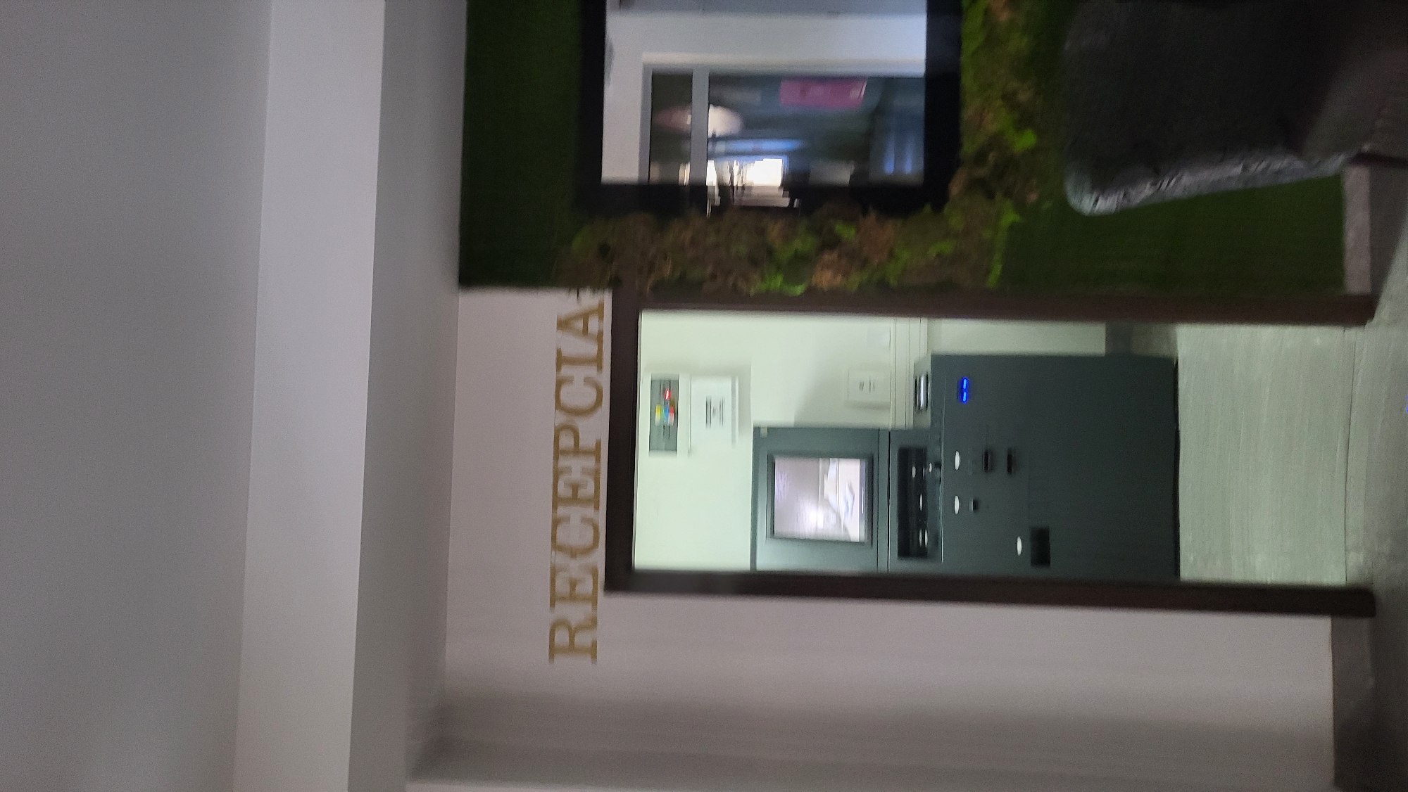 A wide shot, slightly tilted upward, captures an indoor scene with golden lettering on a white wall spelling "RECEPCIA" above a doorway. Through the doorway, a grey ATM machine is visible, with its screen displaying an image. A black chair with a textured surface is partially visible to the right of the doorway, next to a dark rectangular frame with a mossy green interior. The floor appears to be light gray tile. The overall setting suggests a reception area, possibly in a commercial establishment in Prešov, Slovakia, due to the word "recepcia," which is Slovak for reception.