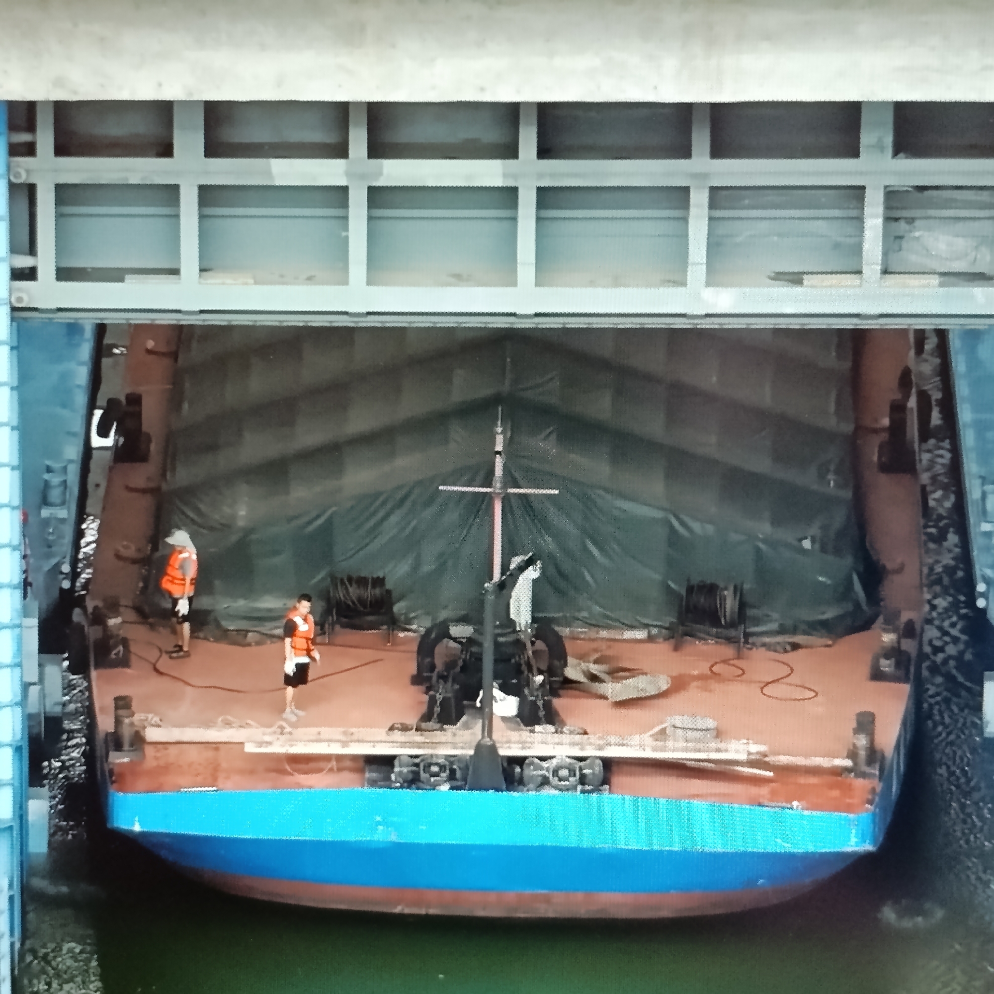 An aerial view shows a barge inside a lock. The barge has a blue and red hull and is covered with a dark green tarp. Two individuals wearing orange life vests are on the deck of the barge. One person, wearing a hat, stands near the port side of the barge. The other person, wearing a white shirt and black shorts, stands closer to the starboard side. A mast with a crossbar stands upright in the center of the barge. Various equipment, including winches and what appears to be a crane, are visible on the deck. The lock walls are visible on the sides and top of the frame, with a metal structure overhead. The barge is situated in greenish water. The scene is located in the Dubăsari town council, Moldova.