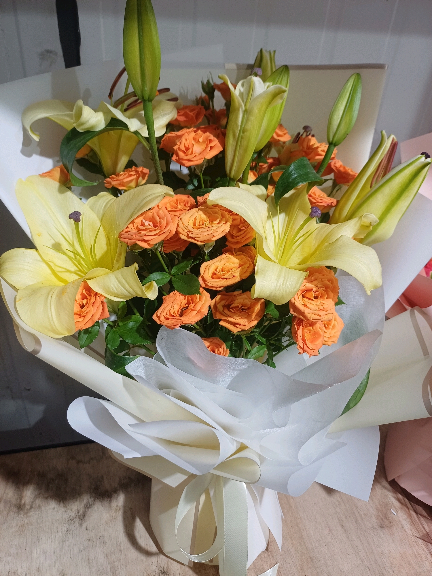 The image shows a close-up, eye-level view of a bouquet of flowers. The bouquet is predominantly composed of large, pale yellow lilies with prominent stamens and numerous small, vibrant orange roses. The flowers are arranged in a dense cluster and are partially wrapped in white and cream-colored tissue paper, with a cream-colored ribbon tied around the base of the bouquet. The background is out of focus, showing a plain, light-colored wall. The flowers are presented on a wooden surface, which appears to be a table or countertop. There is no indication of people, actions, or interactions in the image. The setting does not provide any specific clues to identify a location in Kuala Lumpur, Malaysia, or any other specific geographical place. The image focuses solely on the floral arrangement.