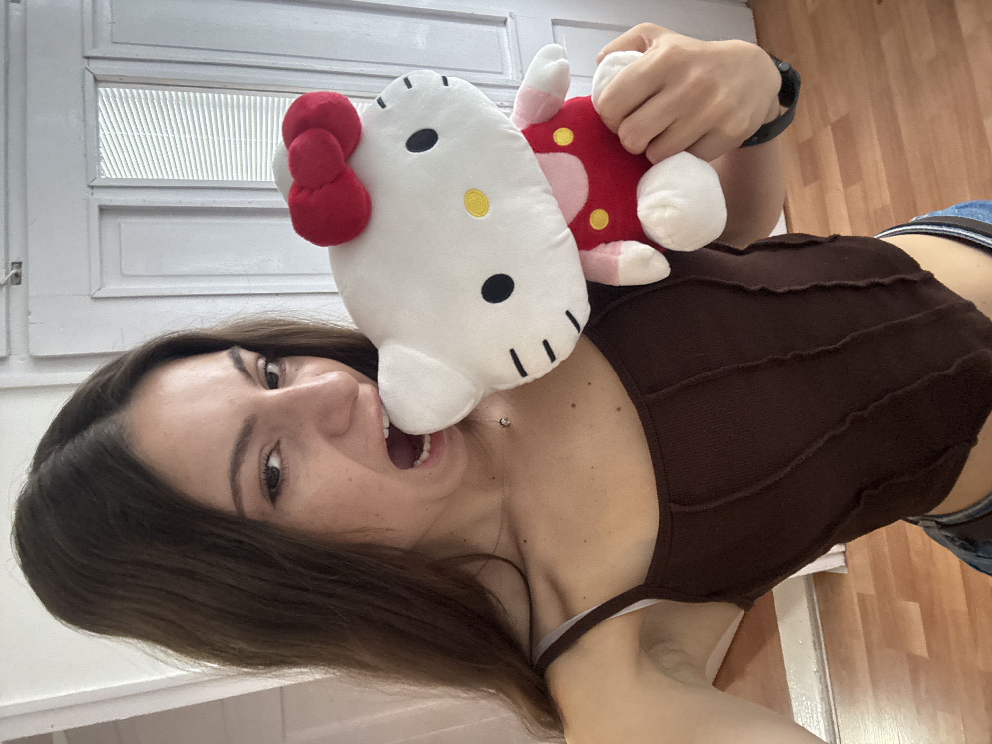 A woman is holding a Hello Kitty plush toy and biting it. The woman is wearing a brown tank top and has dark hair. The background includes a white door with blinds. The location is not discernible as Kaspichan, Bulgaria from the image.