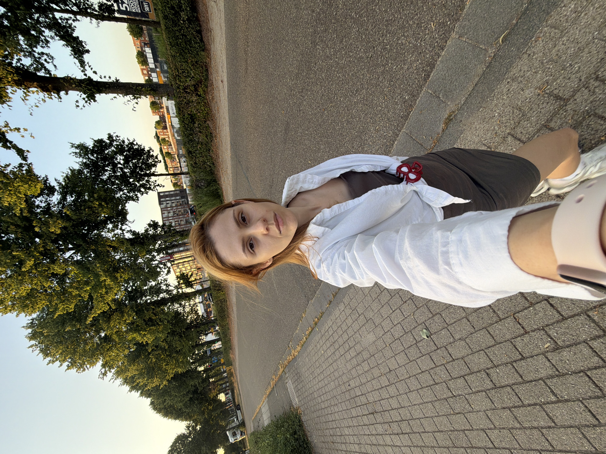 A young woman with shoulder-length blonde hair is taking a selfie while walking on a paved sidewalk next to a road. She is wearing a white open button-down shirt over a dark brown dress and white sneakers. A red hair clip is attached to her dress. The sidewalk is made of grey rectangular pavers. The road is asphalt and appears to be a city street with trees lining the left side. In the background, there are buildings and construction sites, as well as more trees. The sky is clear and light blue with a hint of sunset colors. The image appears to have been taken in Helmond, Netherlands, based on the context.