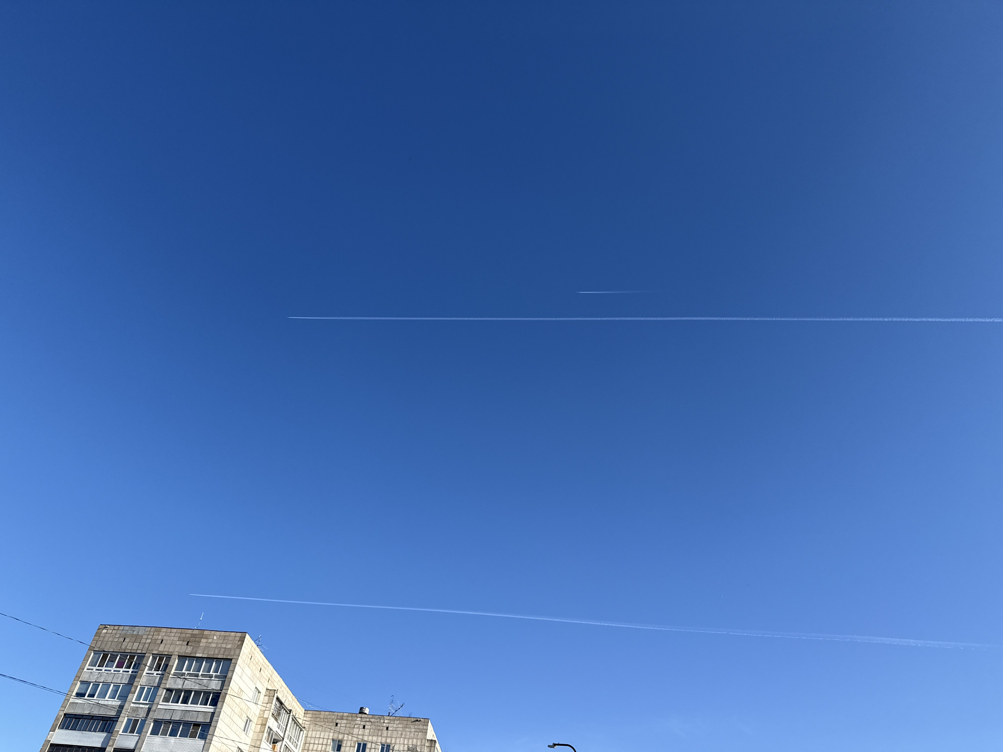 The image shows a clear blue sky over an urban area. In the lower left corner, there is a multi-story building with multiple windows. Two contrails from airplanes stretch across the sky, one higher and one lower. A power line is visible in the lower left of the image.