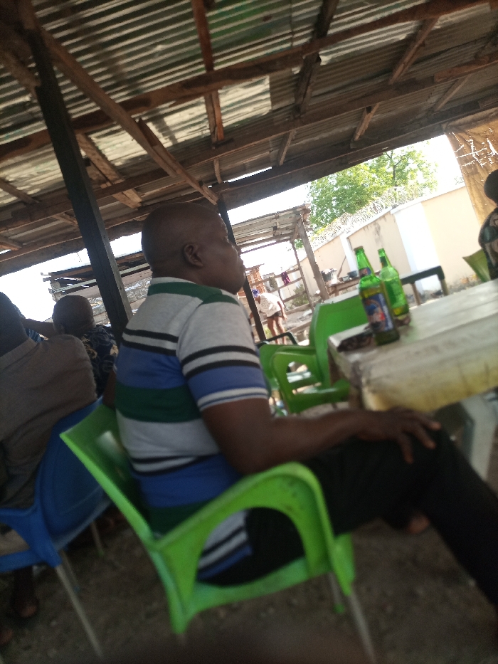 The scene depicts an outdoor or semi-outdoor establishment located in the City of Unknown, Unknown. An adult male with a bald head and dark complexion is seated in profile, facing right, in the foreground. He wears a short-sleeved polo shirt with horizontal stripes in grey, white, green, and blue, and dark trousers. He is positioned on a bright green plastic chair, with his right hand resting on his right thigh. To his right, a light-colored, rectangular table holds two green glass bottles, one with a visible label. Another person is partially visible to the left, seated on a blue plastic chair, wearing a light brown garment. Further left, a third person's head is partially visible. The overhead structure consists of a corrugated metal roof supported by wooden beams and dark vertical posts. The ground appears to be dirt or gravel. In the background, a white wall topped with barbed wire is visible, beyond which are green trees and other distant, indistinct structures. The environment is illuminated by natural daylight.