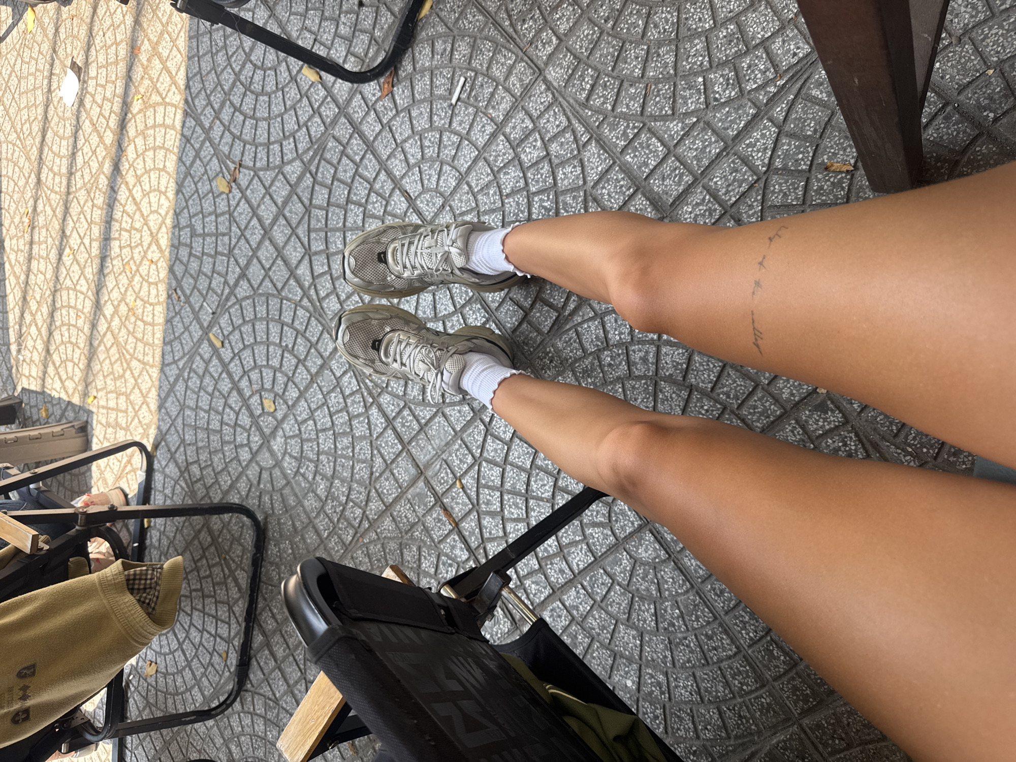 The image presents a top-down view of a person seated in an outdoor setting, most likely in Đà Nẵng, Vietnam, suggested by the typical pavement. The person is wearing athletic shoes with white socks. The lower legs are visible, with a tattoo on the right ankle. The scene includes parts of chairs, and the pavement shows a textured pattern of circular and diamond shapes.