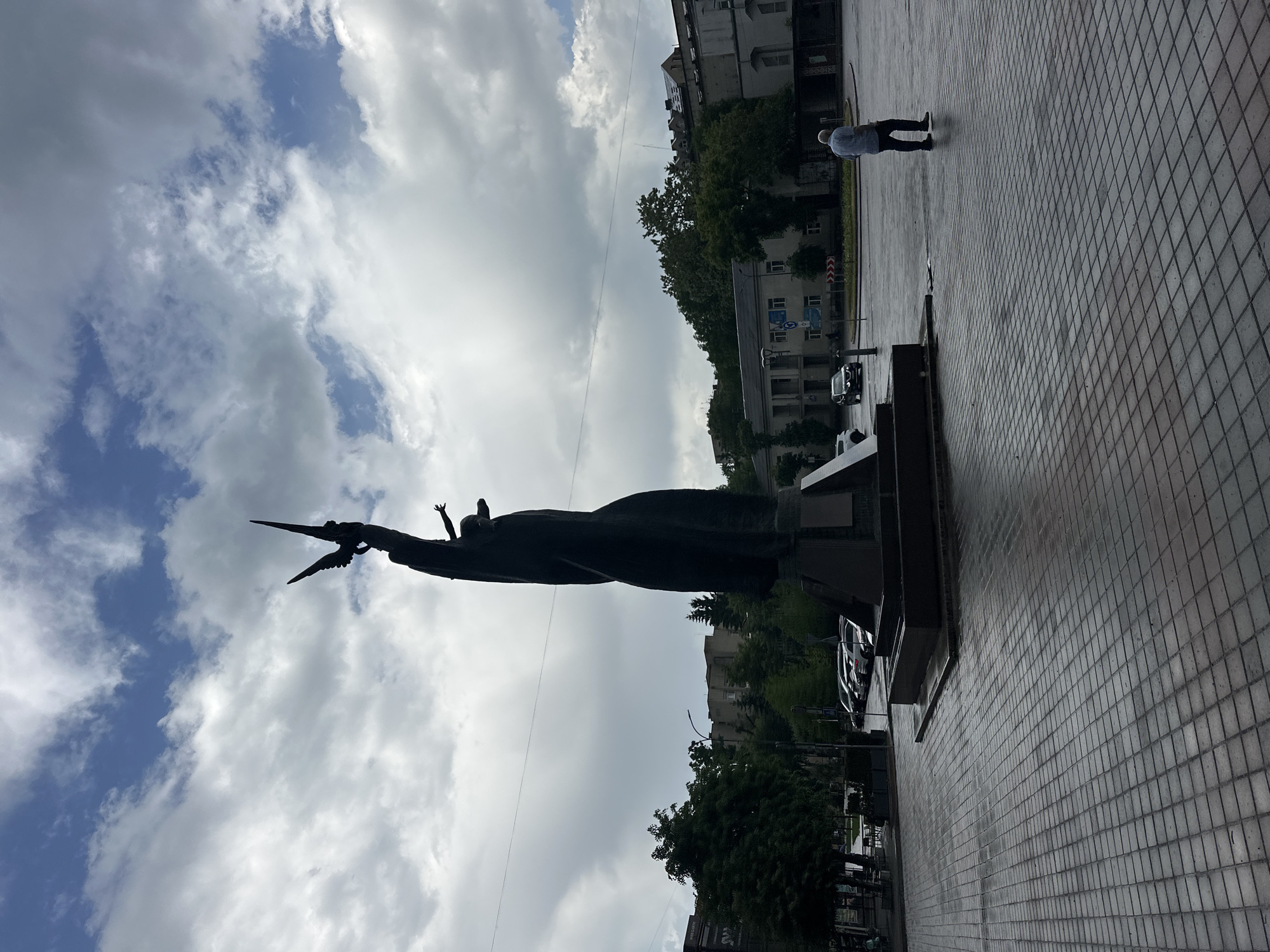The image shows the Monument to the Victims of Communism in Ternopil, Ukraine. The statue depicts a figure holding a dove, likely symbolizing peace. The figure is silhouetted against a cloudy sky. A man is walking on the paved ground near the monument. Buildings and trees are visible in the background. Cars are parked alongside the street. The statue and the environment around it are clear and well-defined.
