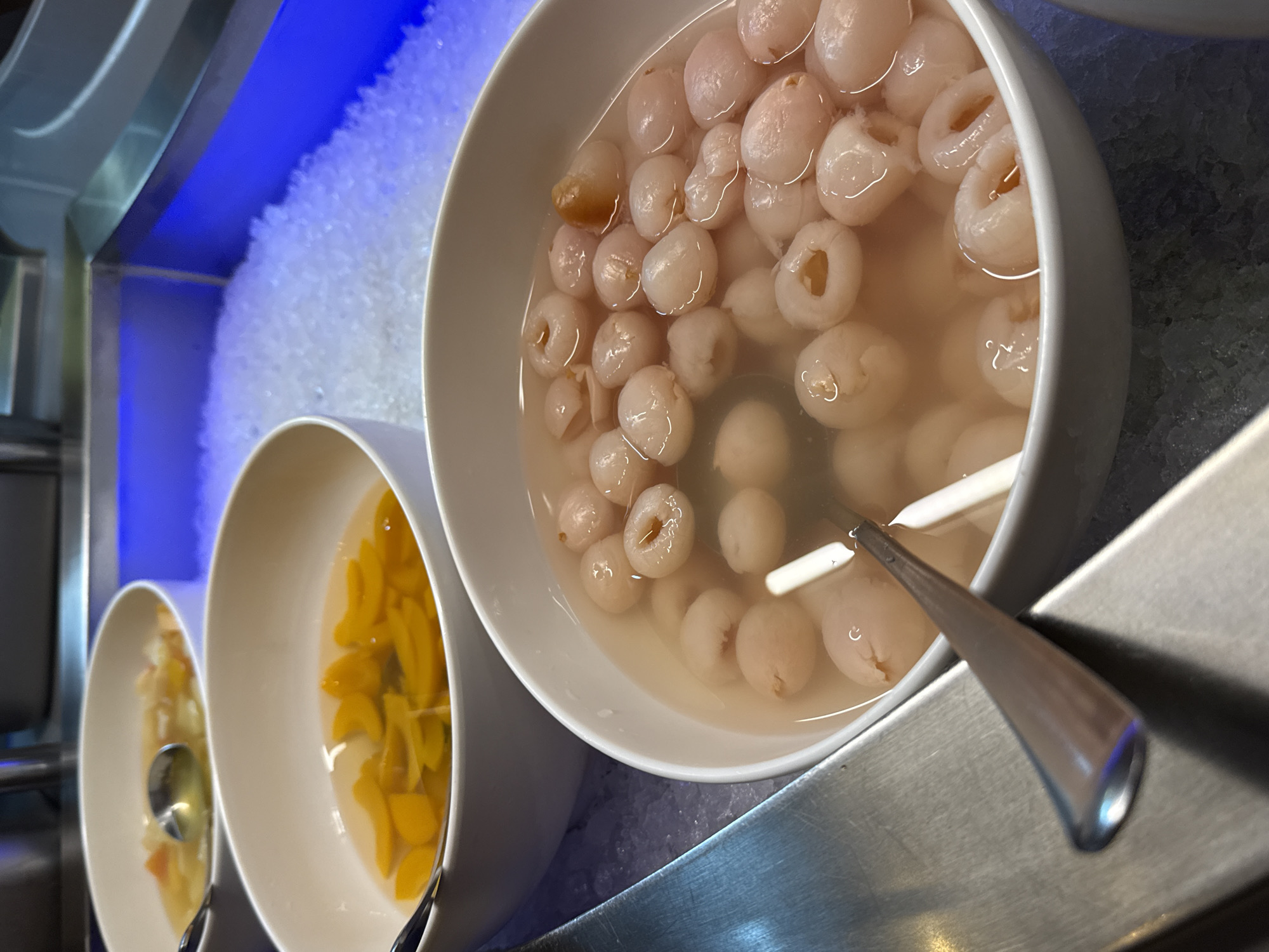 This is a close-up, eye-level, outdoor shot of a buffet with several bowls of fruit salad. In the foreground, a white bowl filled with lychees in syrup is prominently displayed. A silver spoon rests in the lychees. Behind this bowl, another white bowl contains sliced peaches in syrup, with a spoon visible. Both bowls are placed on a stainless steel counter next to a display filled with crushed ice and illuminated by blue lighting. The buffet setting suggests a food service establishment in Nordhausen, Germany.
