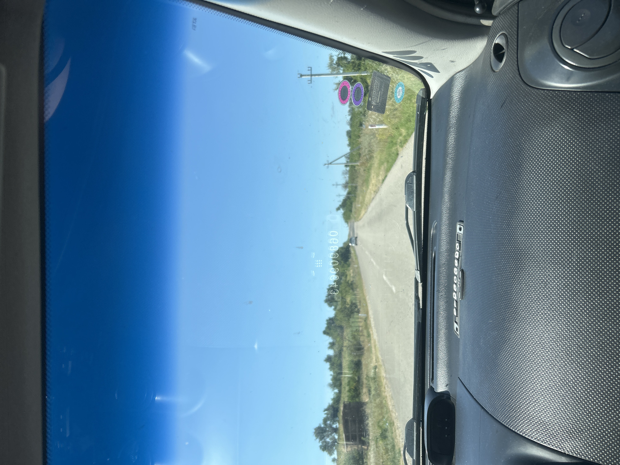 A view from inside a vehicle looking out through the windshield at a paved road on a clear, sunny day. The road stretches into the distance, flanked by dry grass, sparse trees, and utility poles. In the foreground, the dashboard of the vehicle is visible, along with the windshield wipers. There are a few stickers on the windshield, including two circular ones with pink and purple rings and a rectangular one with text. The sky is a vibrant blue.