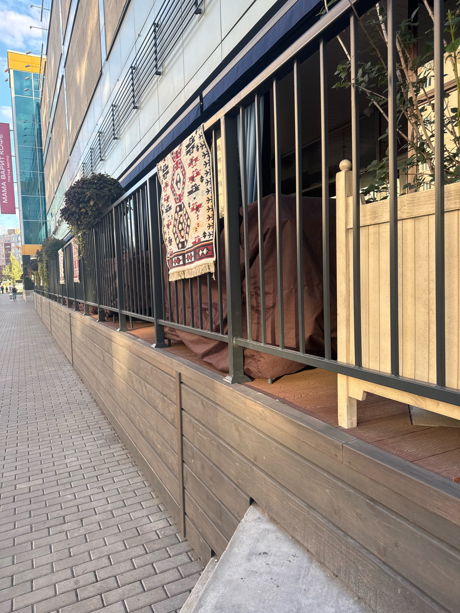 An outdoor seating area of a cafe is depicted, located on a paved walkway in front of a modern building with large glass windows. The seating area is separated from the walkway by a dark metal railing and a wooden barrier. A rug with a geometric pattern is hanging on the railing. Several large brown tarps are draped over what appear to be outdoor furniture. A potted plant is visible on the railing, and a wooden planter box stands to the right. In the background, the facade of the building displays a banner with Russian text, likely indicating the name of the establishment. The walkway is paved with grey rectangular pavers. A few pedestrians are visible in the distance. The lighting suggests it is daytime, with shadows indicating the sun's position. The overall scene is one of an urban commercial environment.
