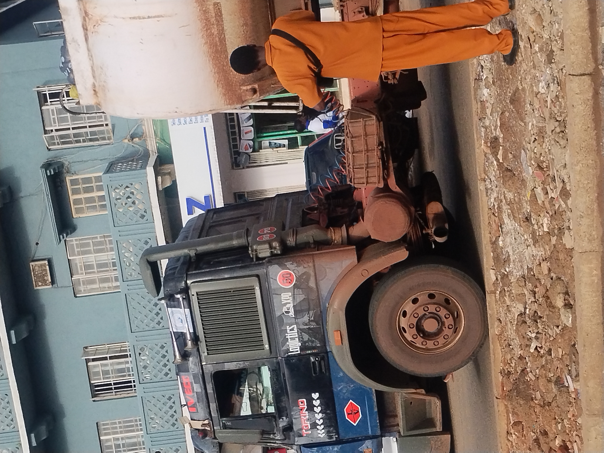 The image captures a scene in the city of Unknown, Unknown, depicting a large truck parked on a road alongside a building with multiple windows. A person in an orange jumpsuit is positioned near the truck's front, seemingly engaged in some kind of activity. The truck is blue and black and labeled with "Logistics XPD" with the number "50". The road surface appears to be dirty and littered with debris.