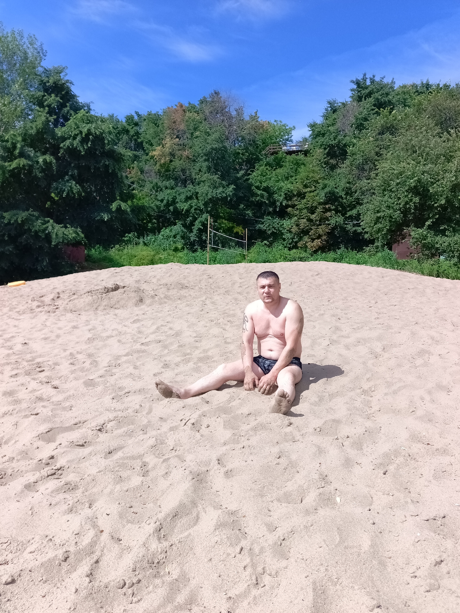 A middle-aged man with short, dark hair and a tattoo on his left arm sits on a sandy embankment. He is shirtless, wearing dark swimming trunks, and his feet are covered in sand. The embankment is part of a beach, with the sea or a large body of water implied by the context. Behind the man, a line of dense green trees forms a backdrop, with a bright blue sky with wispy white clouds overhead. A volleyball net is visible among the trees. The scene suggests a warm, sunny day at a recreational area, potentially within or near the village of Vyazovka, Russia, given the geographical context.