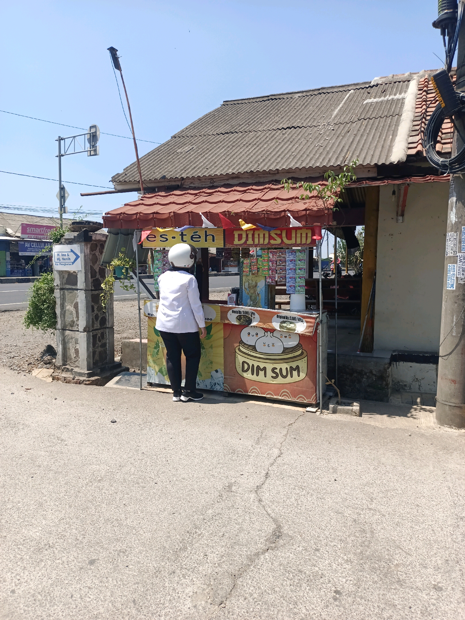 In Subang, Indonesia, a person wearing a white shirt, black pants, and a white helmet stands at a small roadside food stall. The stall displays "es-teh" (iced tea) and "DIM SUM" signage. Several packets of snacks are visible behind the counter. The stall is situated on a paved road, with a stone pillar and greenery to its left, and a utility pole to its right. A traffic light and a building with a corrugated roof are visible in the background. The weather appears sunny with a clear blue sky.
