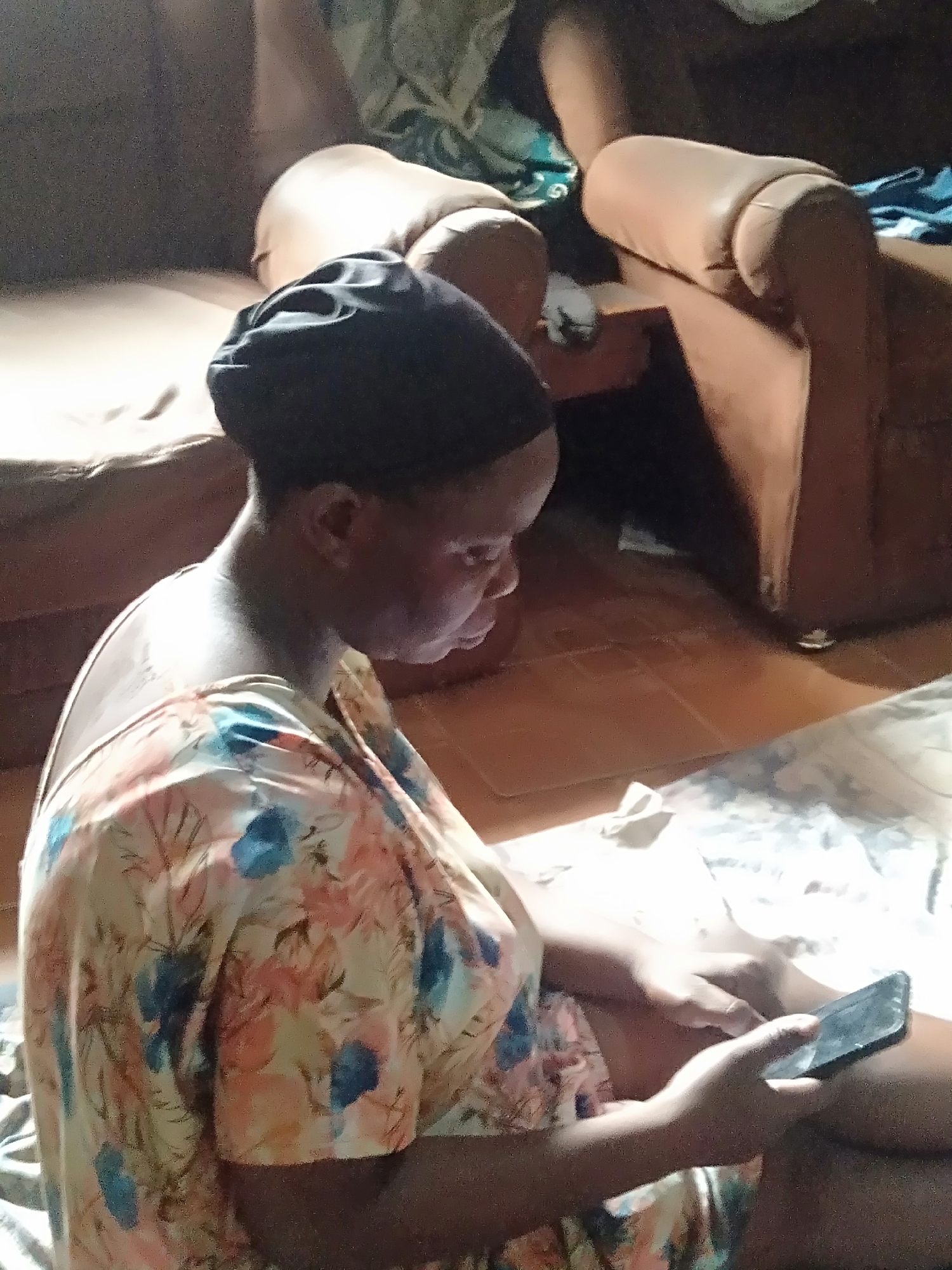 A woman wearing a black head covering and a floral-patterned dress sits on the floor in a domestic interior, looking down at a smartphone she holds in her hands. Behind her is a brown leather sofa. The floor is tiled. The location is Shangisha, Nigeria.