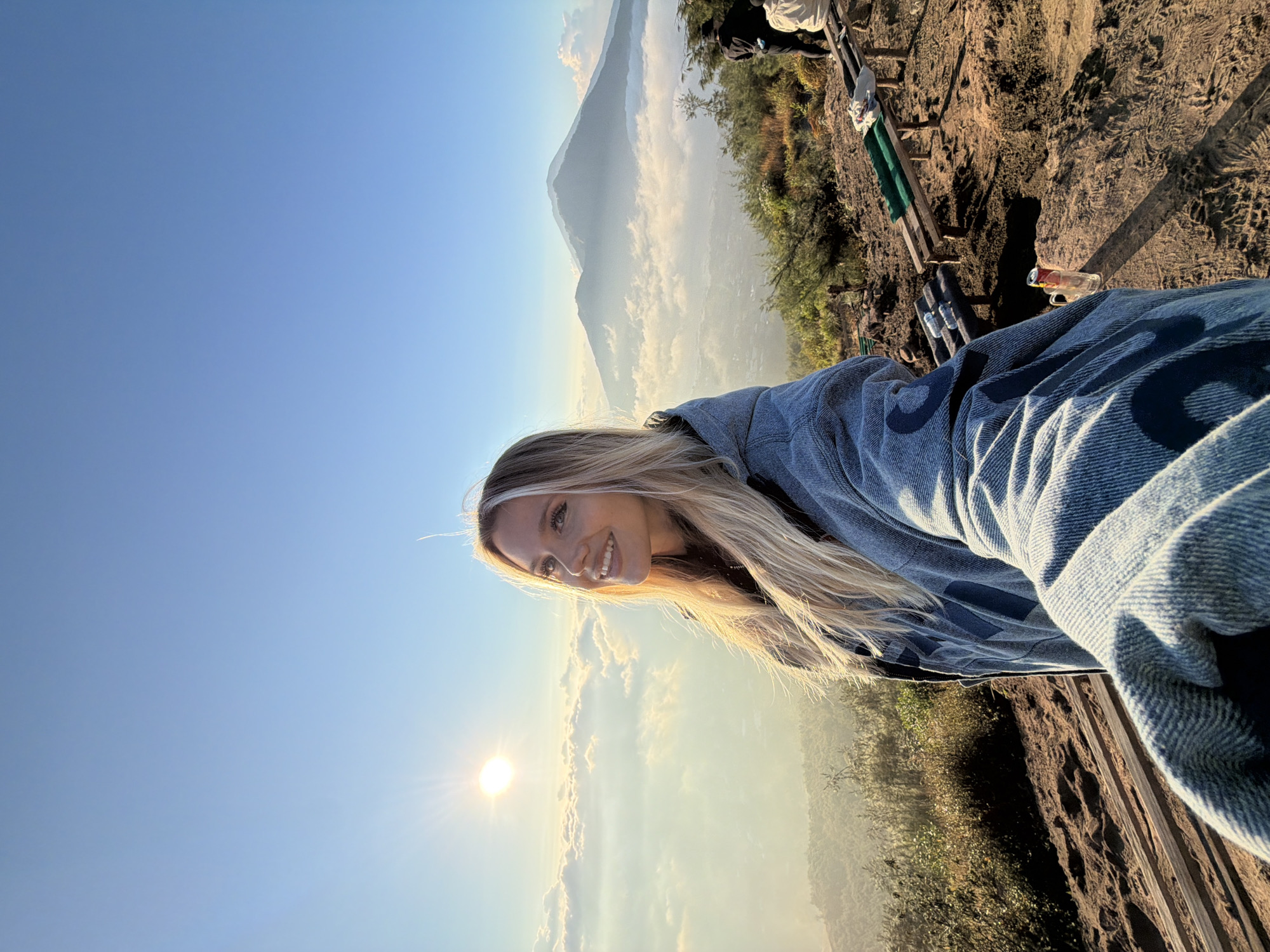 A woman with blonde hair takes a selfie on a mountaintop in Kintamani, Indonesia. She is smiling and looking at the camera, wearing a grey jacket with blue lettering. Behind her, a majestic volcano rises from a sea of clouds, bathed in the warm light of the rising sun. In the foreground, wooden benches are set up for visitors, and a can of drink rests on a rock. The overall scene conveys a sense of peace and natural beauty.