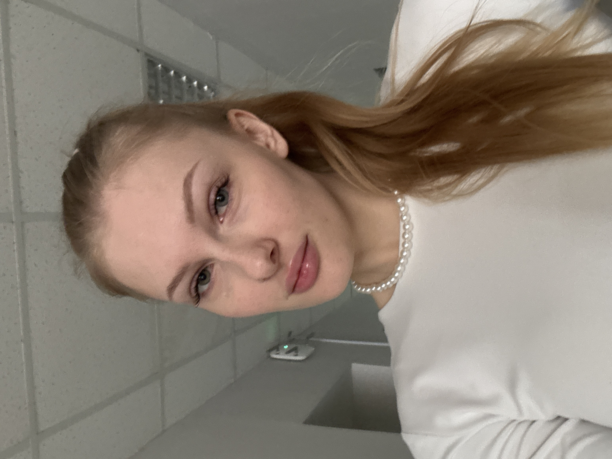 The image is a portrait of a young woman, captured indoors and presented with a significant counter-clockwise tilt. She has fair skin, light-colored eyes (appearing green or blue-green), and long, light brown or blonde hair that flows over her right shoulder. Her hair is partially styled away from her face. She wears natural-looking makeup, highlighting her eyebrows, eyelashes, and lips with a pinkish-nude gloss. Around her neck, she sports a white pearl choker necklace, complementing her simple white, long-sleeved top or dress, visible on her right side.
The woman looks directly at the viewer with a calm, gentle expression.
The background features a light-colored acoustic tile ceiling with a grid pattern, and a plain, light grey wall behind her. Below and to her right, a darker grey surface holds a small, white electronic device with a prominent green indicator light, possibly a router. The setting appears to be an interior space, likely a public or institutional building in Perm, Russia, given the background elements and provided context. No visible text is present in the image.