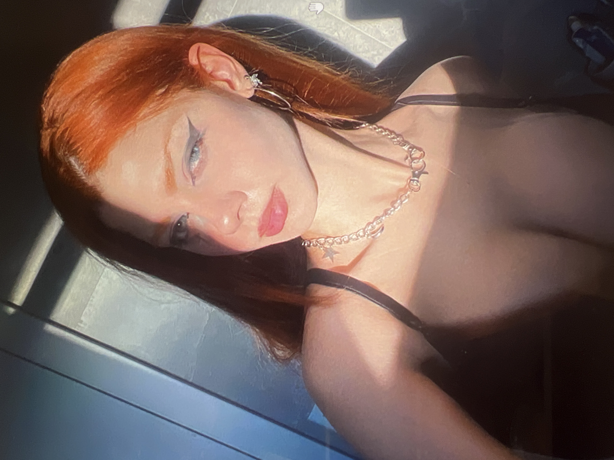 The image is a close-up, self-portrait of a young woman with auburn hair, pale skin, and blue eyes. She is wearing heavy black eyeliner with a winged effect and has bright pink lipstick. A silver chain necklace with star and ring charms is visible around her neck, along with hoop earrings. She is wearing a dark, strappy garment. The lighting suggests it is either early morning or late afternoon, with strong directional sunlight casting shadows and highlighting her face and upper body. The background appears to be an indoor space with a textured, grey-blue wall, possibly made of metal or composite material, with vertical lines suggesting panels or blinds. There is no indication of the city of Minsk, Belarus, or any specific location within it.