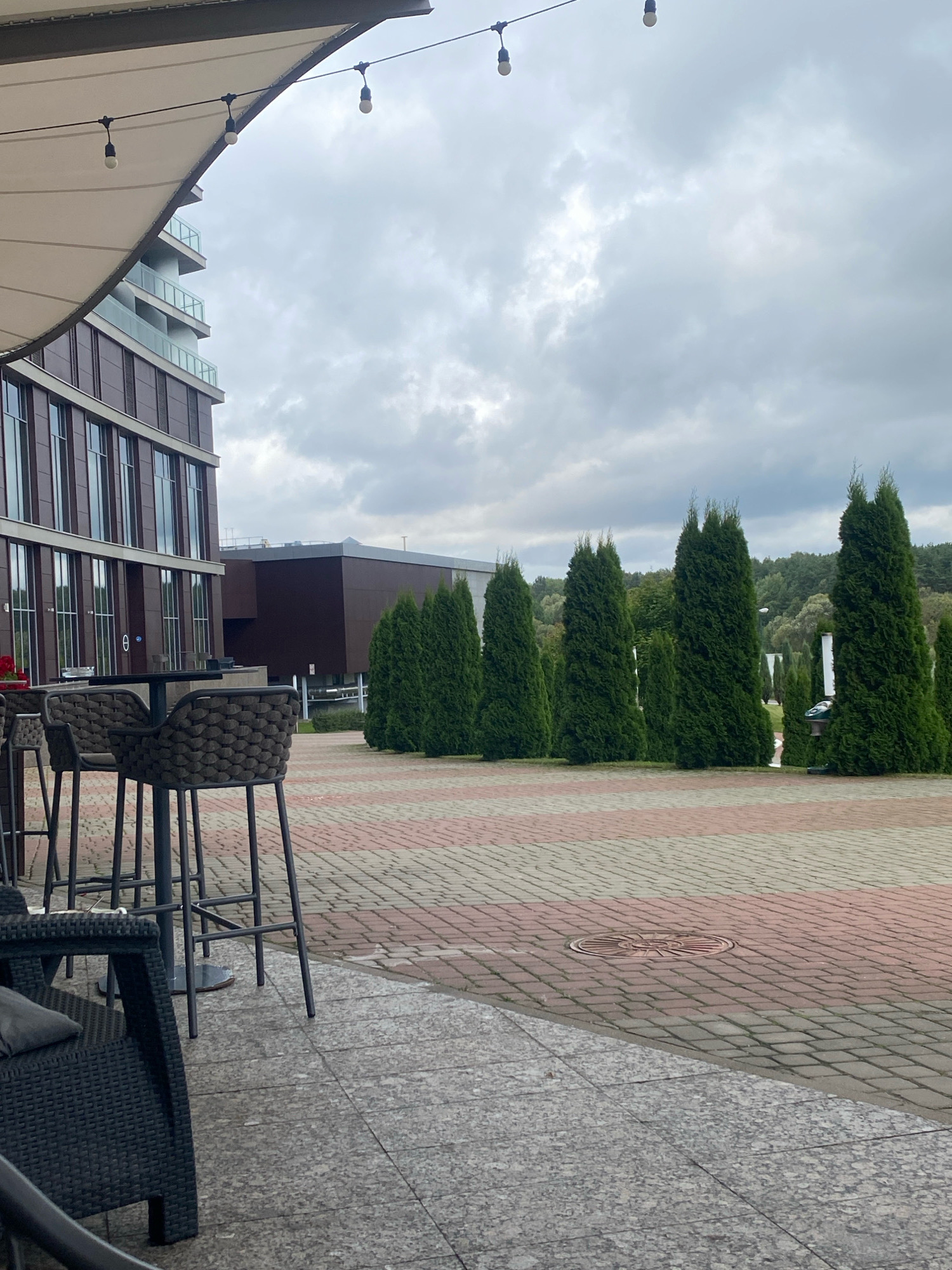 This is a medium-shot, outdoor photograph taken from a slightly low angle, looking towards a modern building and a row of tall, evergreen trees under an overcast sky. In the foreground, a portion of a large, white awning stretches across the top left of the frame, with three exposed light bulbs hanging from a wire beneath it.
To the left, a section of a multi-story building with dark red-brown cladding and numerous large windows is visible. To its right, a smaller, dark brown building is situated behind it. In the middle ground, a paved area with a patterned brick surface, alternating between reddish-brown and light gray, leads towards the trees. Several dark-colored bar stools are positioned around a table in the foreground, suggesting an outdoor seating area. A dark wicker chair is also visible in the lower left corner.
A row of tall, slender, dark green evergreen trees forms a prominent line extending from the middle ground into the background. Beyond the trees, a dense line of natural foliage and trees covers rolling hills, indicating a landscape with natural elements. The sky is filled with heavy, gray clouds.
The scene appears to be an outdoor commercial or public space, possibly a restaurant patio or a promenade adjacent to a modern building complex. Based on the visual cues, this location is likely in Minsk, Belarus, given the architectural style and landscaping. There are no people visible in the image. The overall atmosphere is subdued due to the cloudy weather.