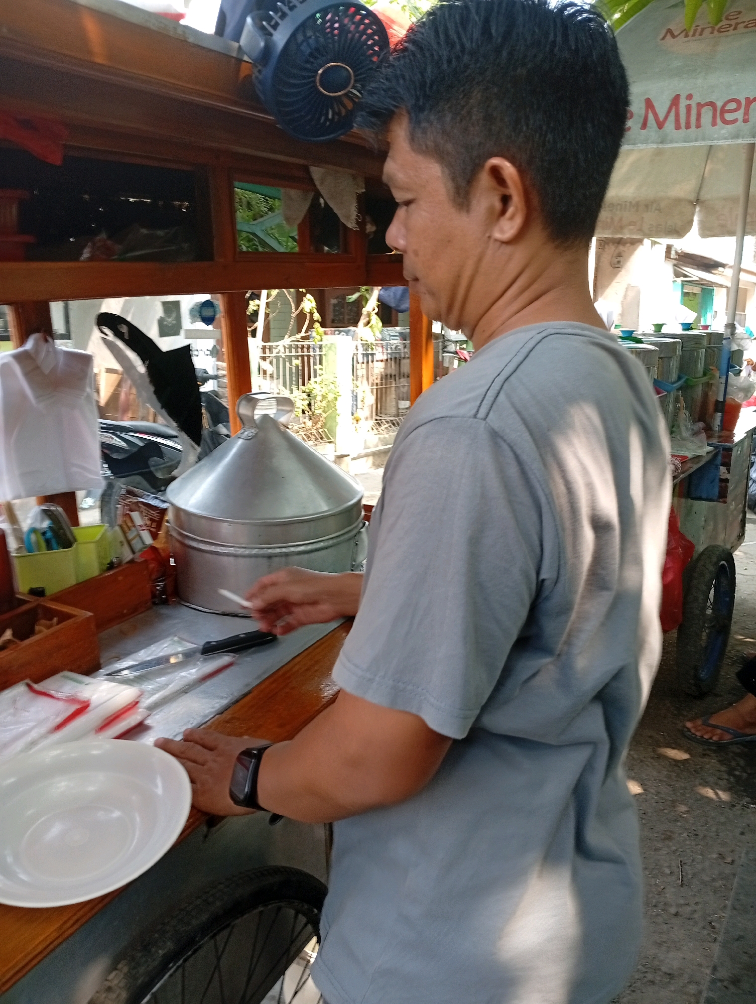 In East Jakarta, Indonesia, a man wearing a gray t-shirt and a black wristwatch stands behind a wooden cart laden with food preparation items. The cart features a large, silver, conical steamer pot, a white plate, plastic-wrapped packets, and a knife. To the left, a small, blue fan is mounted on the cart's upper section. In the background, a vendor's umbrella with the word "Minera" is visible. The scene appears to be outdoors, with sunlight casting shadows on the ground and the man's t-shirt. A tire of the cart is visible in the lower left corner.