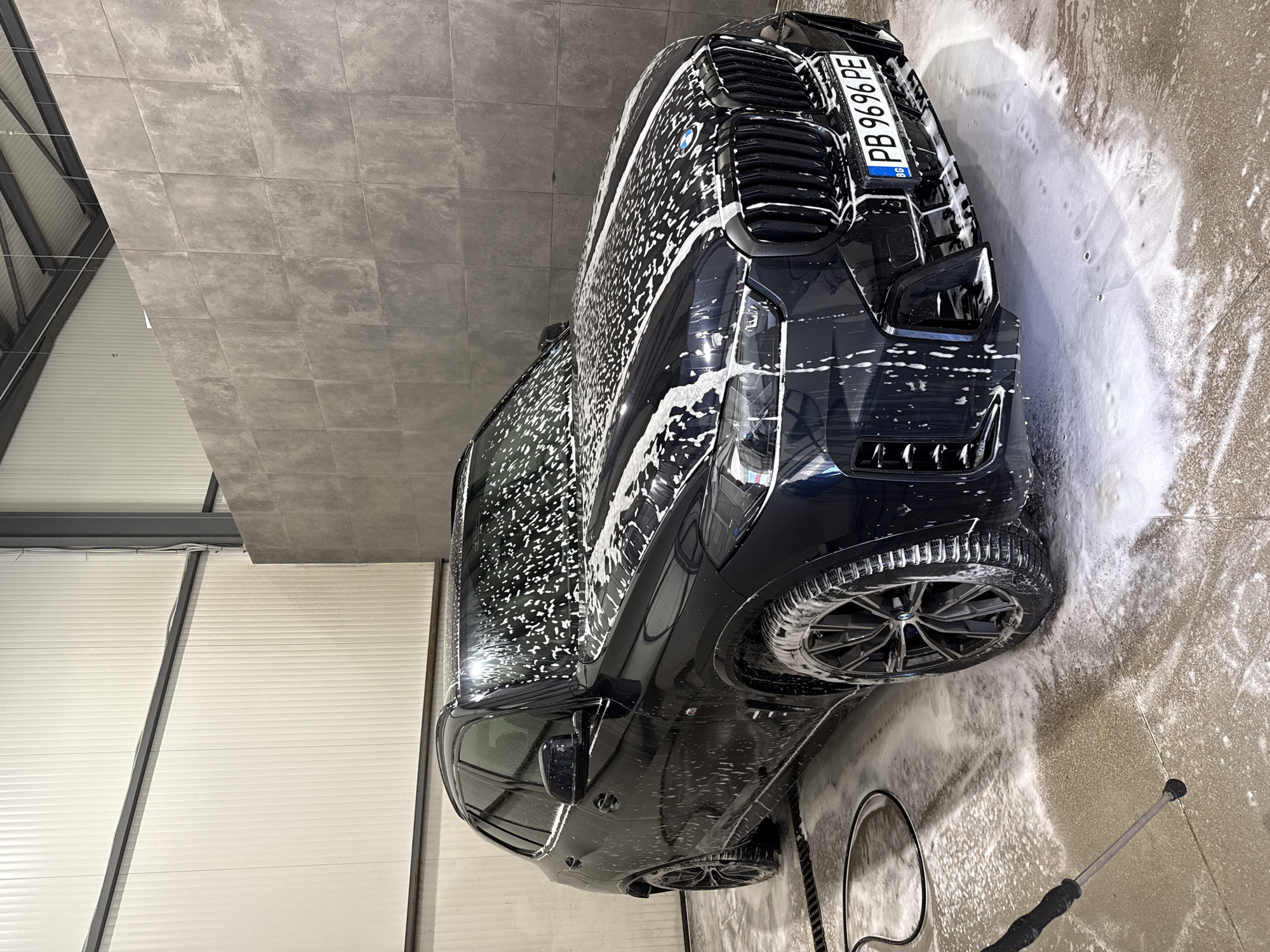 A dark-colored BMW is covered in white soap suds in an enclosed car wash bay. The license plate reads "PB 9696 PE." The car is positioned diagonally within the frame. The walls of the bay are made of grey concrete blocks on one side and white corrugated metal on the other. A power washer wand lies on the floor in the bottom right corner of the frame. The location is in Plovdiv, Bulgaria, indicated by the "PB" on the license plate, which corresponds to the province of Pazardzhik, a region near Plovdiv.