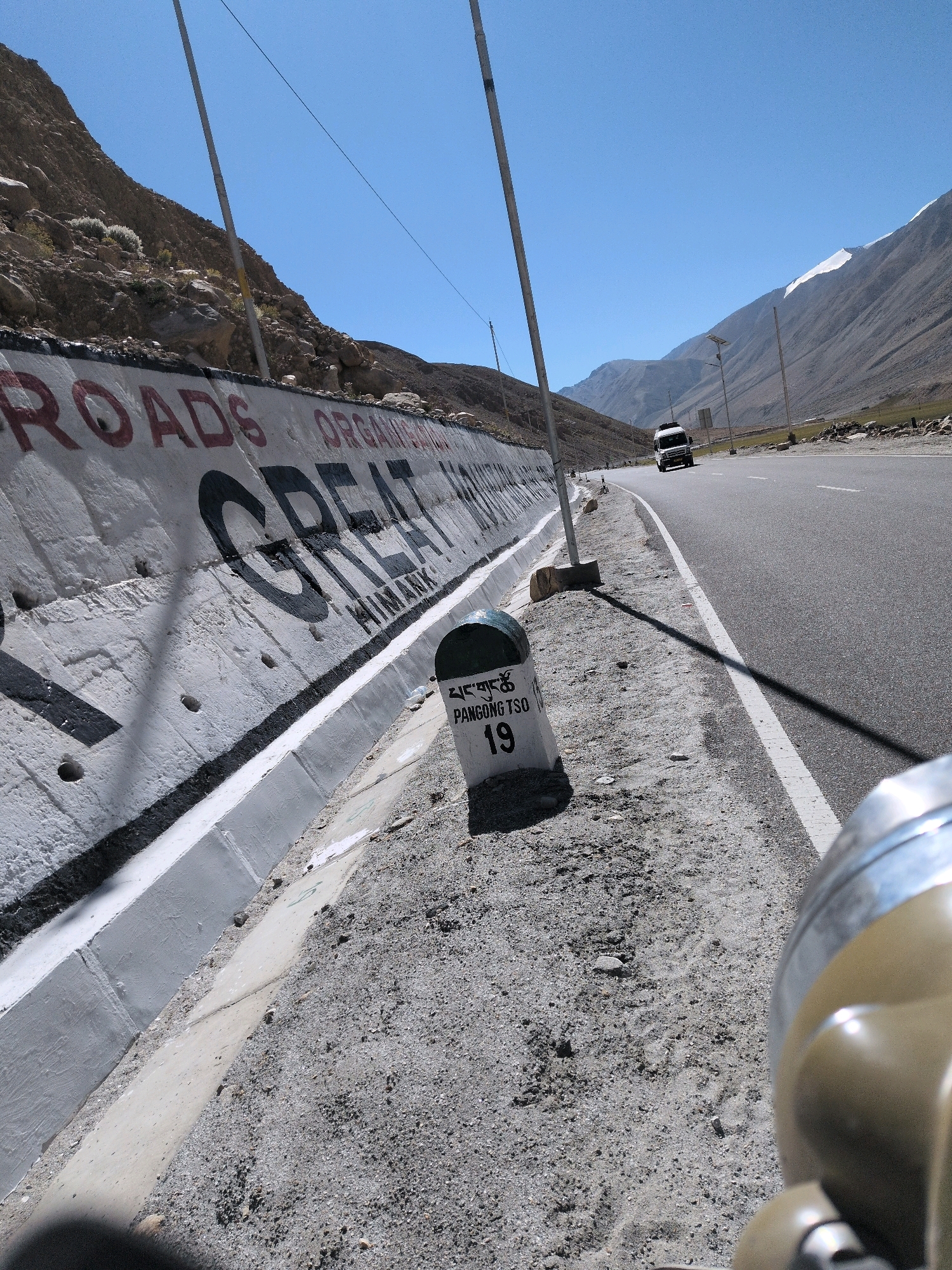 A concrete retaining wall lines the left side of a paved road in a mountainous, arid region. The wall is painted white and features large black lettering that spells out "GREAT HIMALAYAN ROADS ORGANISATION". To the right of the road, a white milestone is visible, marked with Tibetan script and the number "19". The milestone is positioned on a gravelly shoulder next to the road. Further down the road, a white van is traveling away from the camera. The sky is clear and blue, and the surrounding landscape is characterized by barren, rocky mountains and hills. The image appears to be taken from a low angle, possibly from a vehicle.