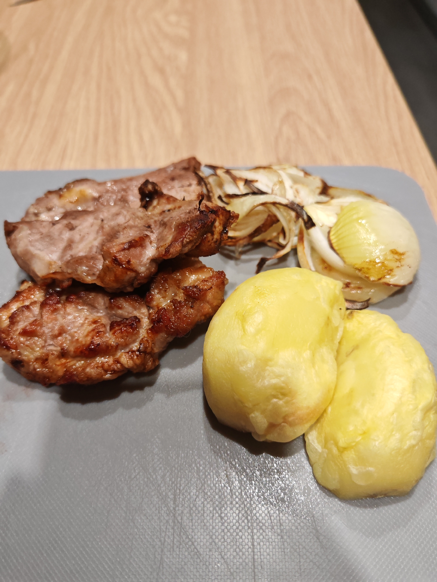 A close-up, eye-level shot shows a meal on a gray cutting board. The meal consists of three pieces of grilled meat, likely pork, with visible grill marks and browning. To the right of the meat are two whole, boiled potatoes, yellow and smooth. Behind the meat and potatoes, there are slices of cooked onion, some caramelized and browned, others translucent. The cutting board rests on a light-colored wooden surface. The overall impression is that of a home-cooked meal. There is no indication of the city of Plovdiv, Bulgaria, or any other specific location.