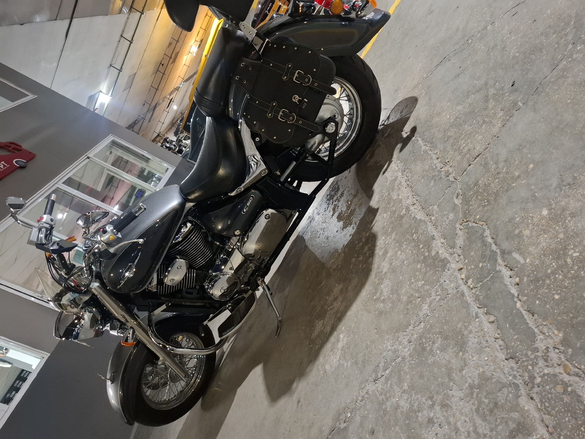 A black cruiser motorcycle is parked on a concrete floor in what appears to be an indoor parking garage. The motorcycle is angled, with the front wheel on the left and the rear wheel on the right. It has chrome accents on the engine, handlebars, and wheels, which are spoked. Black saddlebags are attached to the rear of the motorcycle. The background shows a grey wall with windows and some yellow markings on the floor, characteristic of a parking area. The lighting suggests an artificial light source. No people are present in the image. The image is a medium shot, capturing the motorcycle from a slightly low angle. The overall scene is industrial and utilitarian. The location is identified as Barri De Porta, Spain, though this specific detail is not visually discernible from the image itself.