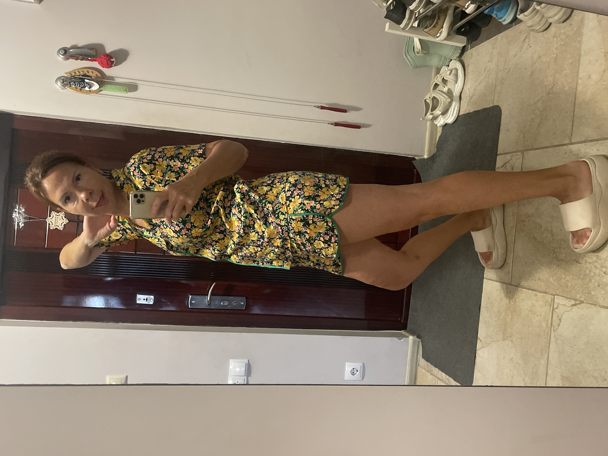 A full-body, mirror selfie shows a woman standing in a hallway. She is wearing a yellow and black floral print pajama set consisting of a short-sleeved top and shorts. The pajama set has a green trim. She is also wearing white platform slides. The woman has her right arm bent at the elbow and her hand on the back of her neck. Her left hand is holding a smartphone, which is angled to capture the image.
The hallway features a dark wooden door with a silver handle and lock to the woman's right. On the wall to her left, there are two light switches and an electrical outlet. The floor is tiled with beige and cream squares. A dark gray mat is on the floor in front of the door. To the far right of the image, a shoe rack is visible, containing various shoes, including sandals, sneakers, and boots. There are also two red jump rope handles hanging from the wall to the right of the woman. The scene appears to be indoors, likely in a residential setting. The user has indicated this image was taken in the city of Sochi, Russia.