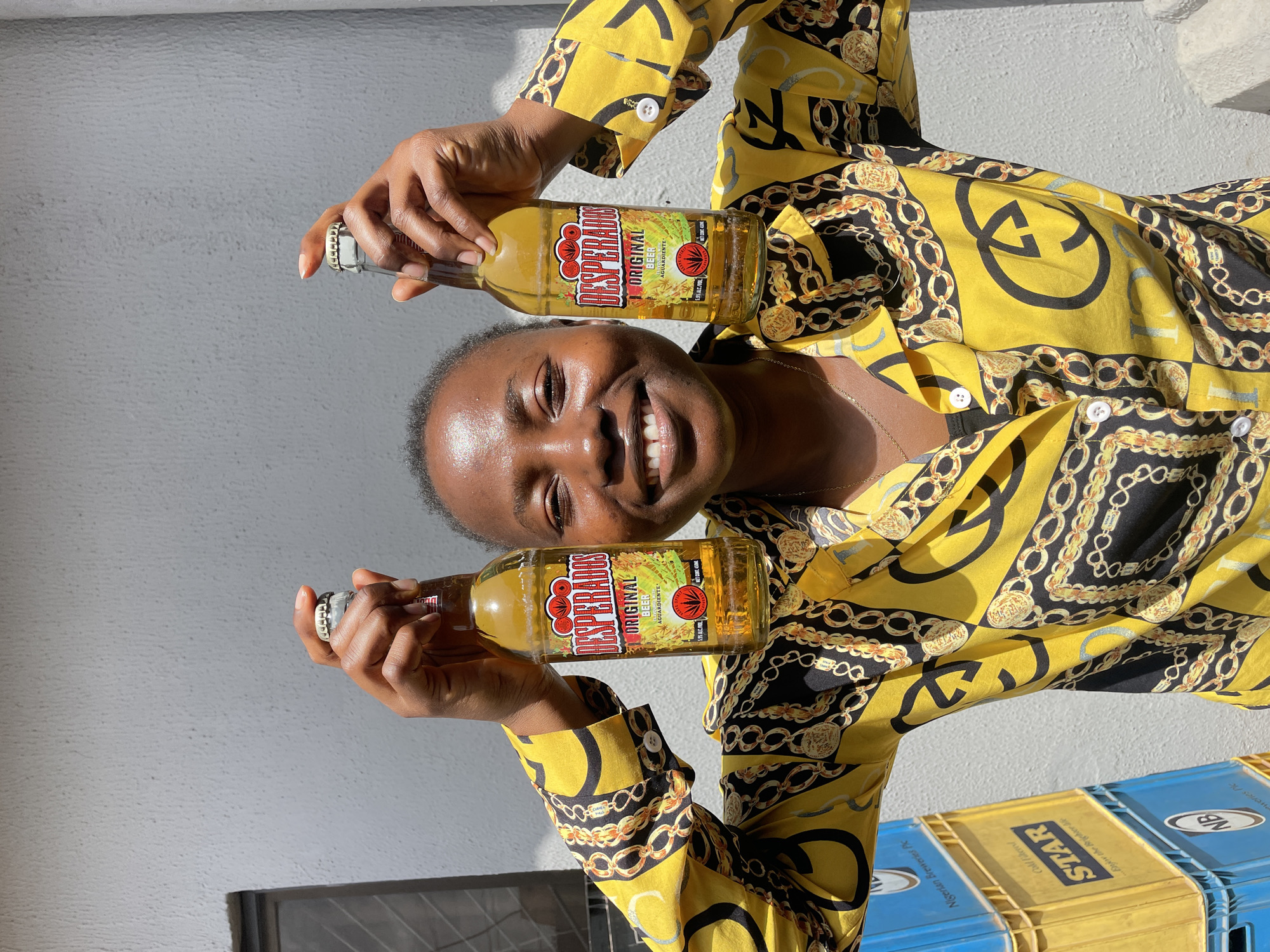 A person, likely a woman based on her hands and jewelry, is shown holding two bottles of Desperados Original beer. She is wearing a yellow shirt with a gold chain and black pattern. Her left hand is holding the top of one bottle, while her right hand is holding the bottom of another. The person is smiling brightly, suggesting happiness or enjoyment. The scene appears to be outdoors, with a light-colored, textured wall in the background and bright sunlight casting shadows. The presence of what looks like beer crates in the lower right corner suggests a commercial or social setting related to beverages. The time of day is likely daytime due to the strong sunlight and shadows. Visible text on the bottles confirms they are "Desperados Original Beer Aguardiente" with an alcohol by volume of 5.9%. The crates also show text including "Nigerian Breweries Plc", "STAR", and "NB". The location context provided is Jos, Nigeria.