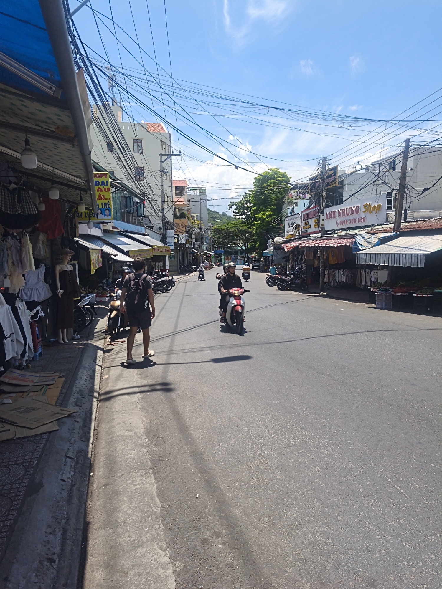 This is a street-level, eye-level, vertical photograph taken on a sunny day in Nha Trang, Vietnam, likely on a commercial street. The image depicts a bustling street scene with shops lining the left side of the road. Several motorcycles are present, with two people riding them actively in the foreground and middle ground. On the sidewalk to the left, there are shops displaying clothing on racks and mannequins. Merchandise is also laid out on cardboard boxes on the ground. The road itself is paved and appears to be in normal condition. Overhead, a complex network of electrical wires is visible against a bright blue sky with scattered clouds. Buildings of varying heights, typical of urban Vietnamese architecture, are visible in the background. The overall impression is one of everyday commerce and street life in a Vietnamese city.
