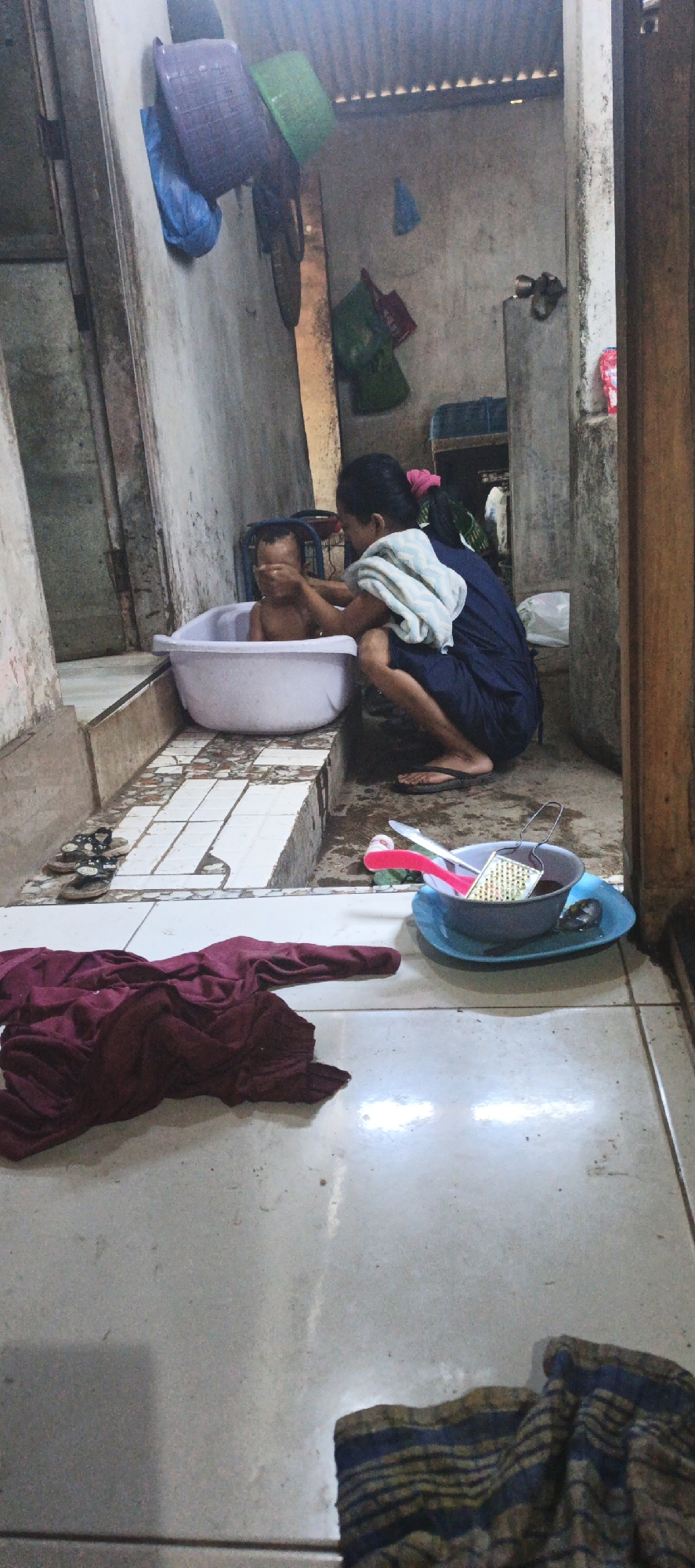 In an outdoor, possibly rural, setting in Kasiyan, Indonesia, a mother is bathing her young child in a white plastic tub. The child is in the tub, and the mother is kneeling beside it, tending to the child. She has a light-colored towel draped over her shoulder and is using it to wash the child. The scene appears to be an informal arrangement, with the bathtub placed on a tiled step outside. In the background, there are some hanging baskets and other household items, suggesting a modest living space. The overall impression is one of domestic care and everyday life. The lighting suggests it might be daytime. There is no visible text in the image.