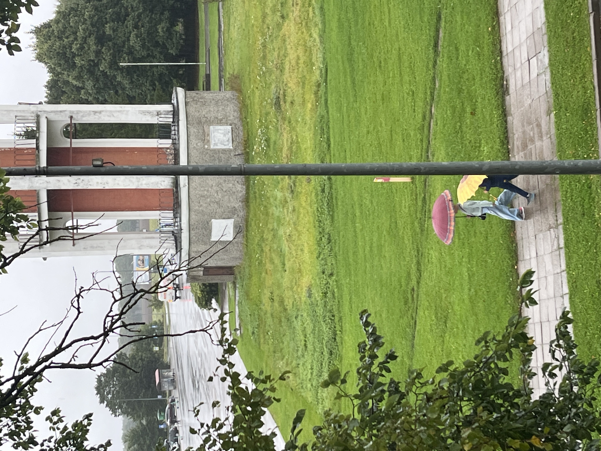 The image depicts a cloudy, rainy day in Kohtla-Järve linn, Estonia. In the foreground, branches of a tree with green leaves partially obscure the view. A paved walkway runs horizontally across the lower portion of the frame. Two individuals are walking on the walkway, one holding a red umbrella and the other a yellow umbrella. The person with the red umbrella is wearing light blue pants and a light grey top. The person with the yellow umbrella is wearing a dark blue outfit. To the right of the walkway is a well-maintained green lawn. Further in the background, there is a tall, cylindrical tower with red and white vertical stripes, and a grey stone base. Beyond the tower, a line of trees forms the backdrop. In the far left of the image, a wet asphalt road is visible with parked cars and a bus stop shelter.