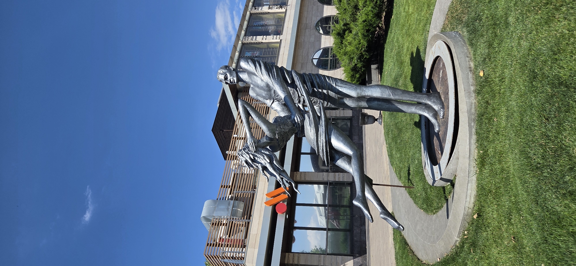 This is a full-frame, vertical, eye-level shot of a metallic sculpture depicting two figures in a dynamic embrace, located in Rostov-on-Don, Russia. The sculpture is positioned outdoors on a circular concrete base surrounded by manicured green grass. A curved concrete pathway leads towards the sculpture from the left.
In the background, there is a modern, multi-story building with a rooftop terrace featuring wooden railings and exposed ductwork. The building's facade is primarily light-colored with large glass windows. A prominent orange and red logo is visible on the building's exterior, likely a commercial establishment. The sky above is a clear, bright blue with a few wispy white clouds. Lush green bushes are planted to the right of the sculpture. The lighting suggests a sunny day.
The sculpture itself features a male figure holding a female figure who is arched backward. Both figures appear to be made of a polished, silver-toned metal. The female figure's hair flows dramatically behind her, and her legs are extended, giving a sense of movement. The male figure's clothing is rendered in a flowing style, as is the female figure's attire. The overall impression of the sculpture is one of passionate movement and unity.