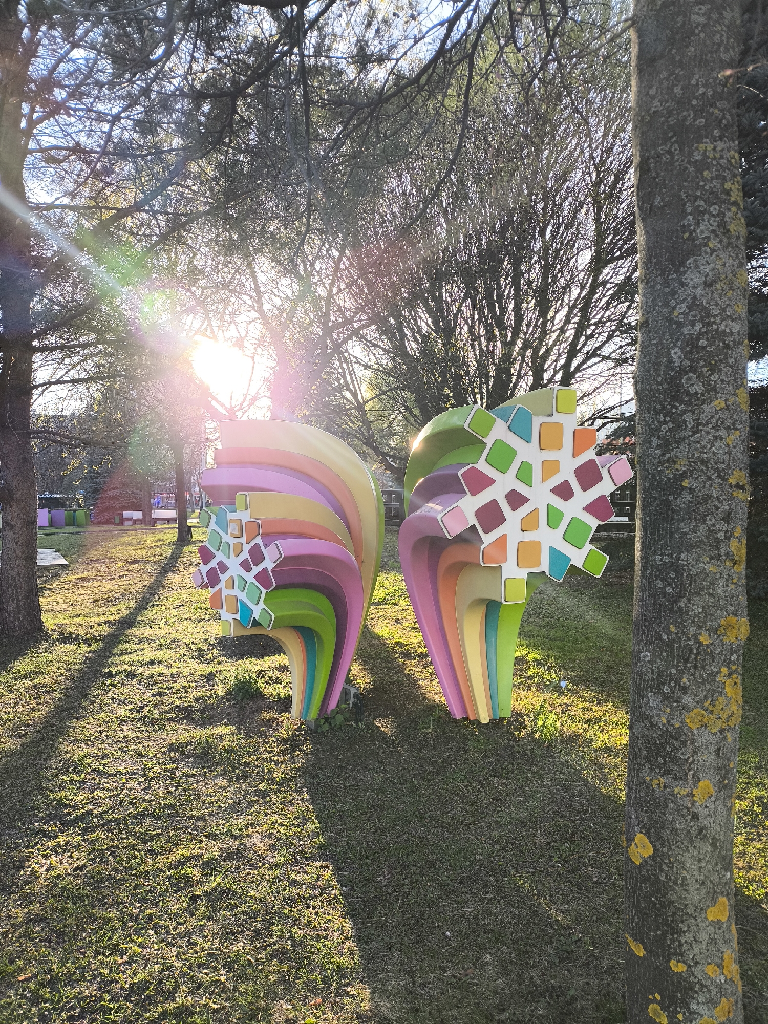 This outdoor image captures two vibrant, abstract sculptures situated in a sunlit park, likely in Moscow, Russia. The main subjects are the two large, colorful installations, resembling stylized cross-sections of organic forms or perhaps abstract representations of large nerve bundles. Each sculpture features a central, snowflake-like pattern composed of various geometric shapes in white, green, orange, pink, red, and blue, surrounded by layered, curved sections in a rainbow of colors including pink, purple, blue, green, yellow, and orange.
The sculptures stand on a grassy patch of ground, which appears somewhat dry in areas, indicating late winter or early spring. Tall trees with mostly bare branches frame the scene, with one tree trunk covered in yellow lichen visible on the right side of the frame.
The strong, low-angle sunlight, indicative of either early morning or late afternoon, shines brightly from behind the trees, creating prominent lens flares and long shadows across the grass. This backlighting silhouettes the tree branches and illuminates parts of the sculptures and the ground with a golden glow. In the background, beyond the sculptures, more trees and what appears to be a colorful outdoor recreational or seating area can be faintly seen.
No people are visible, suggesting a quiet moment in the park. There is no discernible activity taking place other than the static presence of the art installations within the natural environment. No visible text is present in the image. The overall mood conveyed is bright and whimsical due to the colorful sculptures contrasting with the natural, sun-dappled setting.