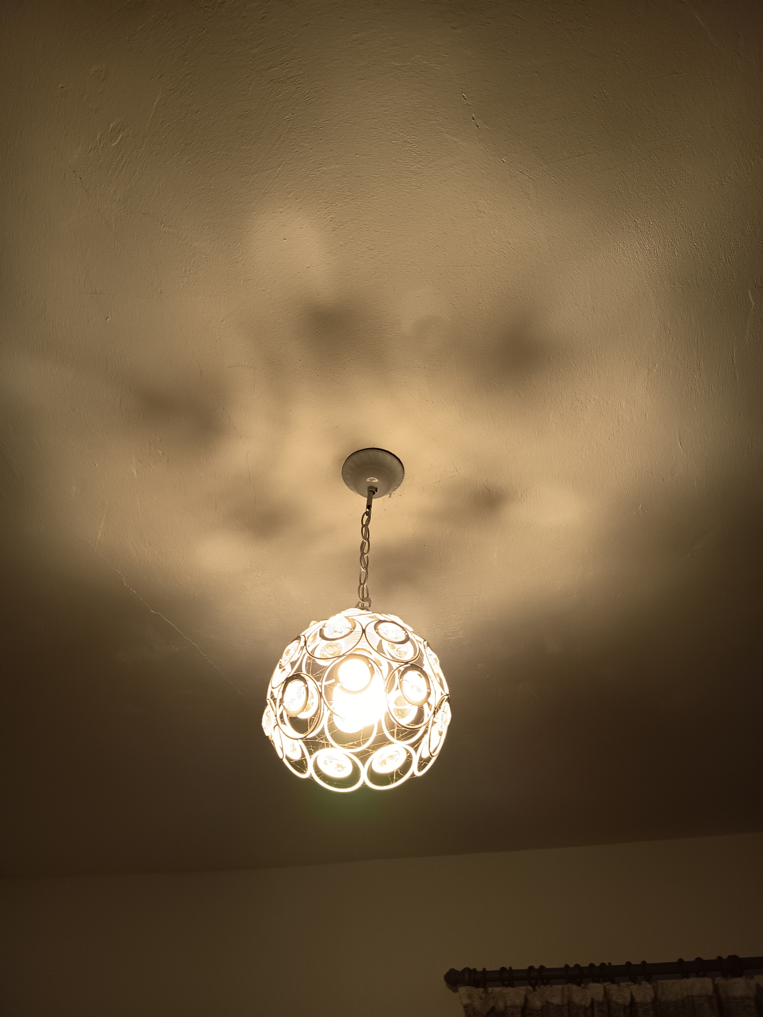A softly lit, interior view shows a round, decorative light fixture suspended from a white base on a textured, off-white ceiling. The ceiling shows subtle shadows cast by the fixture. The lower part of the image is darker, revealing a partially visible window with patterned curtains at the very bottom right. The location is Unknown, Unknown. No people are visible in the scene.