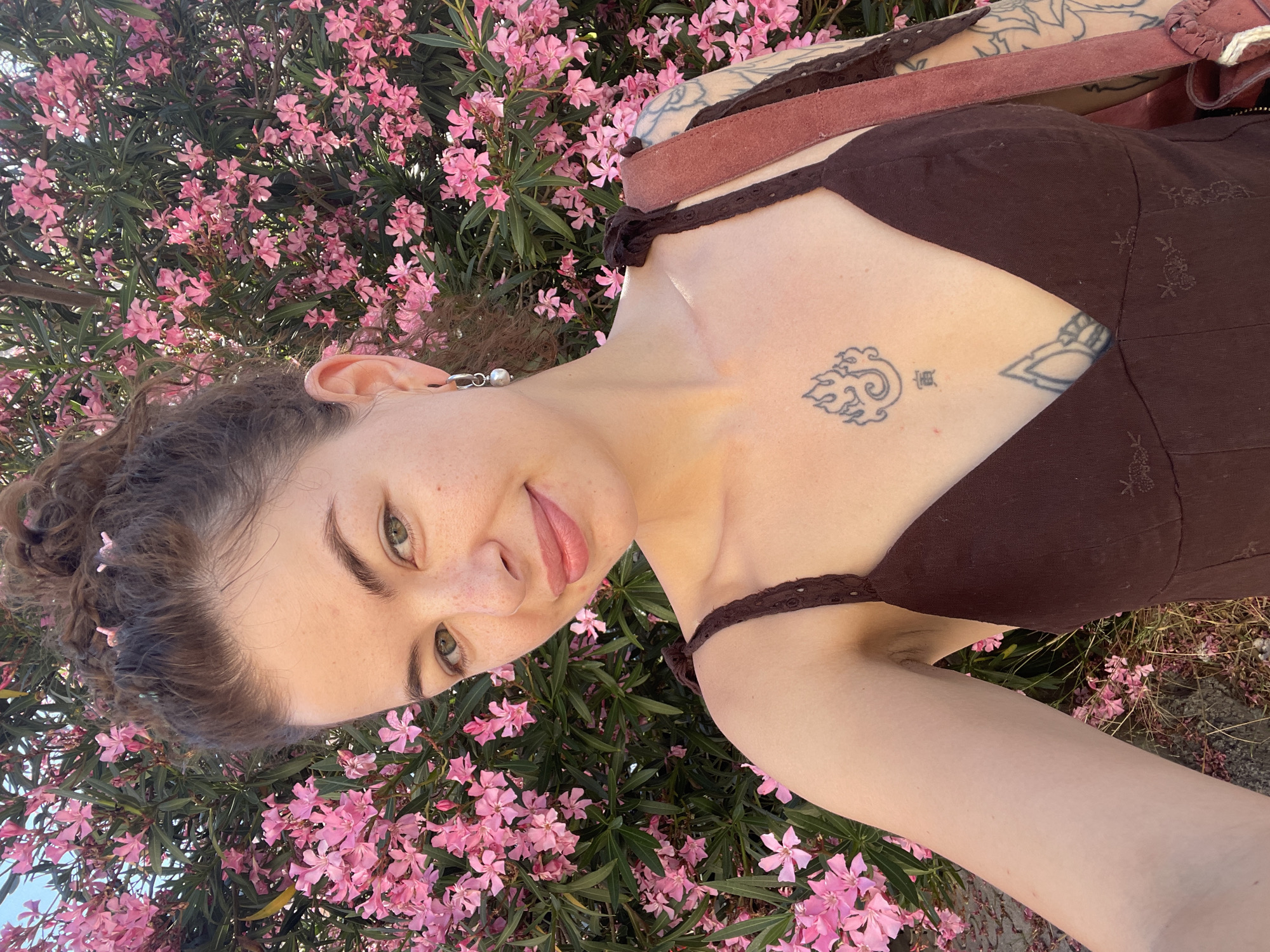 A young woman with curly brown hair pulled into a bun is taking a selfie outdoors in front of a flowering bush with pink blossoms. She is wearing a dark brown dress with thin straps and has a tattoo on her chest. A bag strap is visible over her shoulder. The background is filled with the pink flowers and green leaves of the bush. The lighting suggests it is daytime. The location is the city of Budva, Montenegro.