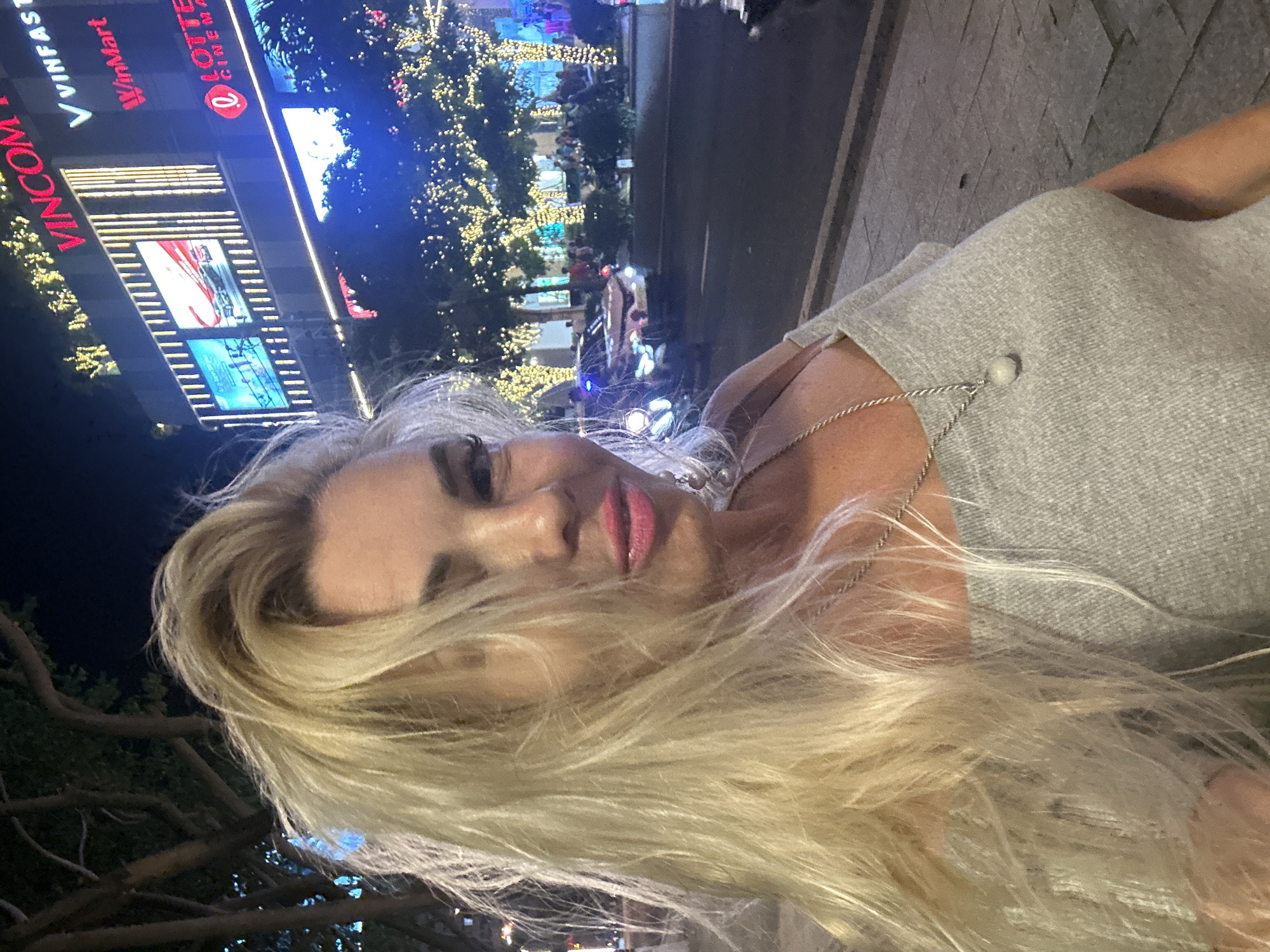 A woman with long blonde hair poses for a selfie at night in Nha Trang, Vietnam. Behind her is a tall building with illuminated signs for "Vincom," "WinMart," and "LOTTE Cinema." Trees decorated with strings of lights are visible in the background, along with a street scene with people and vehicles. The woman is wearing a grey top with a thin strap and a delicate silver necklace. The street appears to be paved with light-colored stone.