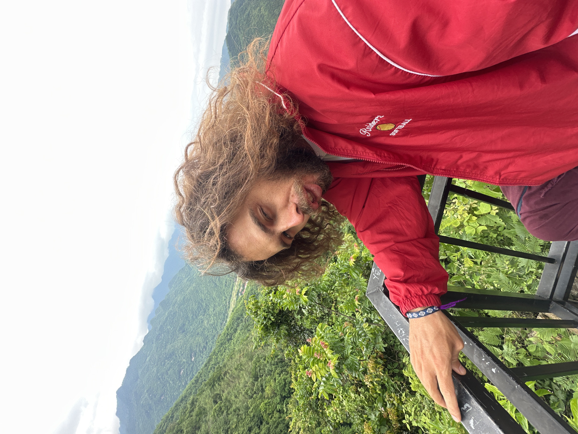 In Mai Chau, Vietnam, a man with long, curly brown hair and a beard stands near a railing overlooking a lush, green mountainous landscape. He is wearing a red jacket with white trim and the text "Raiders Softball" embroidered on it. His right hand rests on the black metal railing, which is marked with graffiti. The background features rolling hills covered in dense vegetation and a cloudy sky. The man is looking down and to his left, with his mouth slightly open.