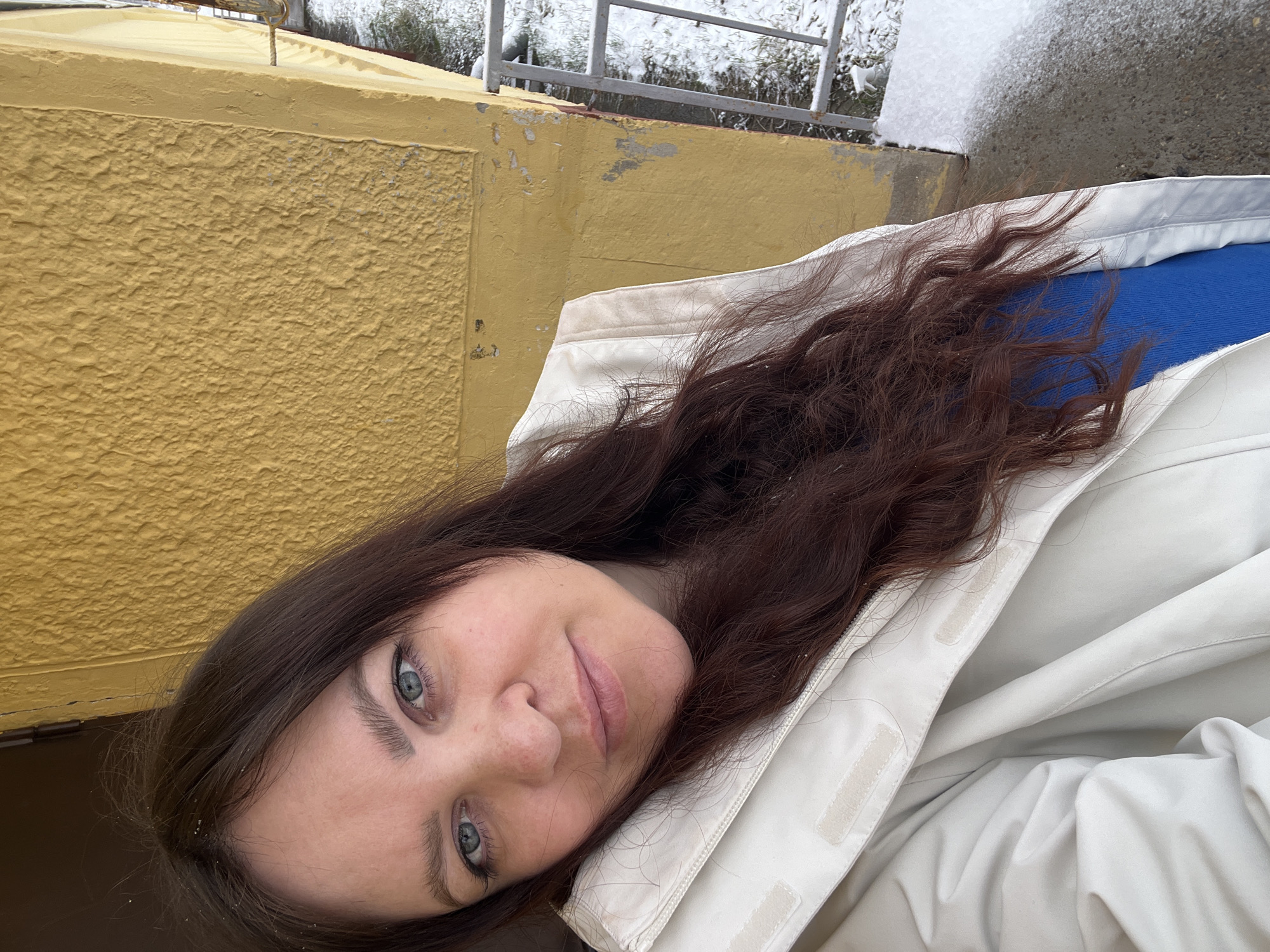 A woman with brown hair and blue eyes lies on a light-colored jacket. She is looking at the camera with a slight smile. Behind her is a yellow, textured wall. In the background, there is a metal railing and snow-covered ground. The location is Noviy, Russia.