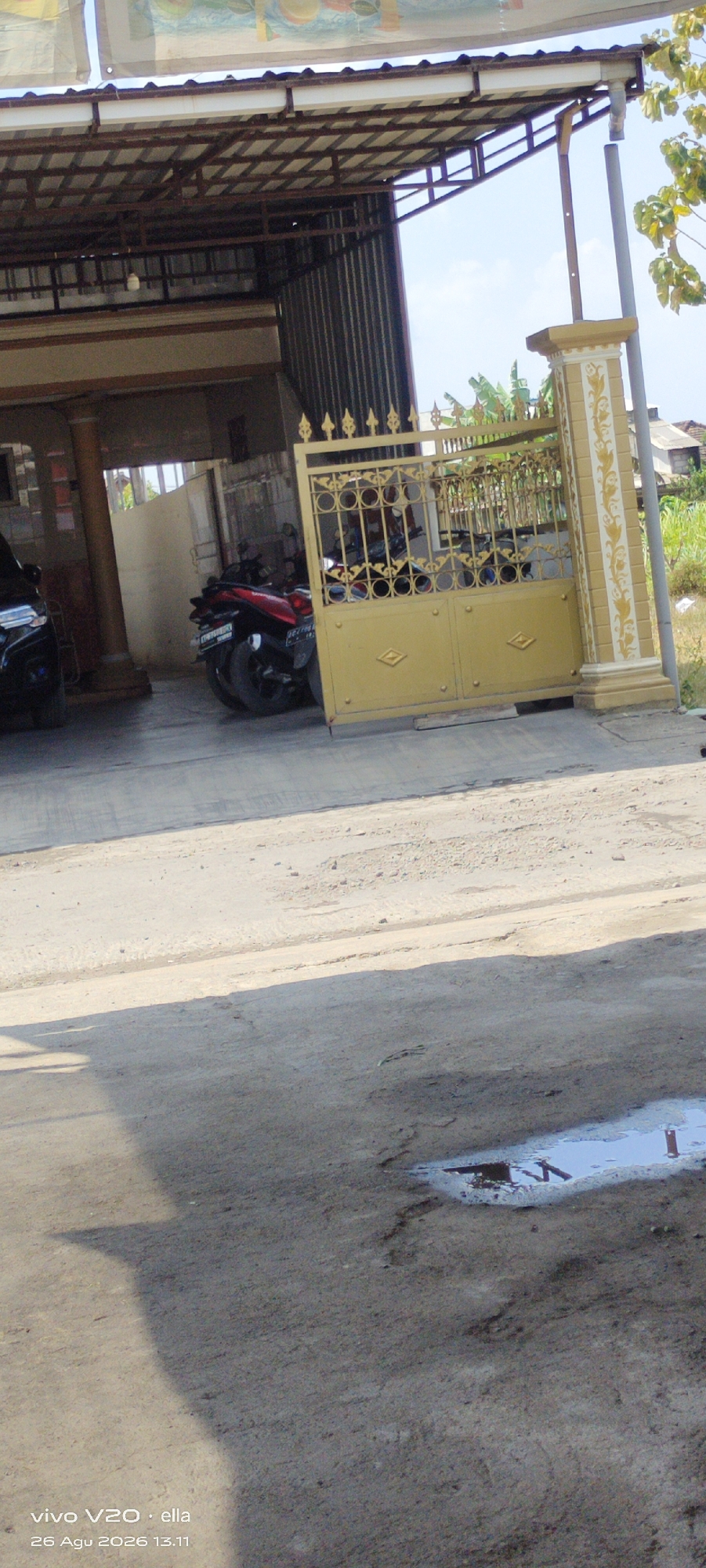 This image depicts an outdoor scene in Simo, Indonesia, featuring a residential or commercial property with a covered carport or building extension. A yellow metal gate with decorative circular patterns and diamond accents is prominently displayed, partially open. Behind the gate, two motorcycles are visible, one red and black, the other partially obscured. A black vehicle is parked to the left of the gate. The ground is paved with a light-colored, somewhat rough surface, showing some patches of shadow. In the foreground, a small puddle of water is present on the pavement. The environment appears to be a sunny day, with a clear sky and some foliage visible in the background. The image is overlaid with a timestamp and device information indicating "vivo V20 · ella 26 Agu 2026 13.11".
