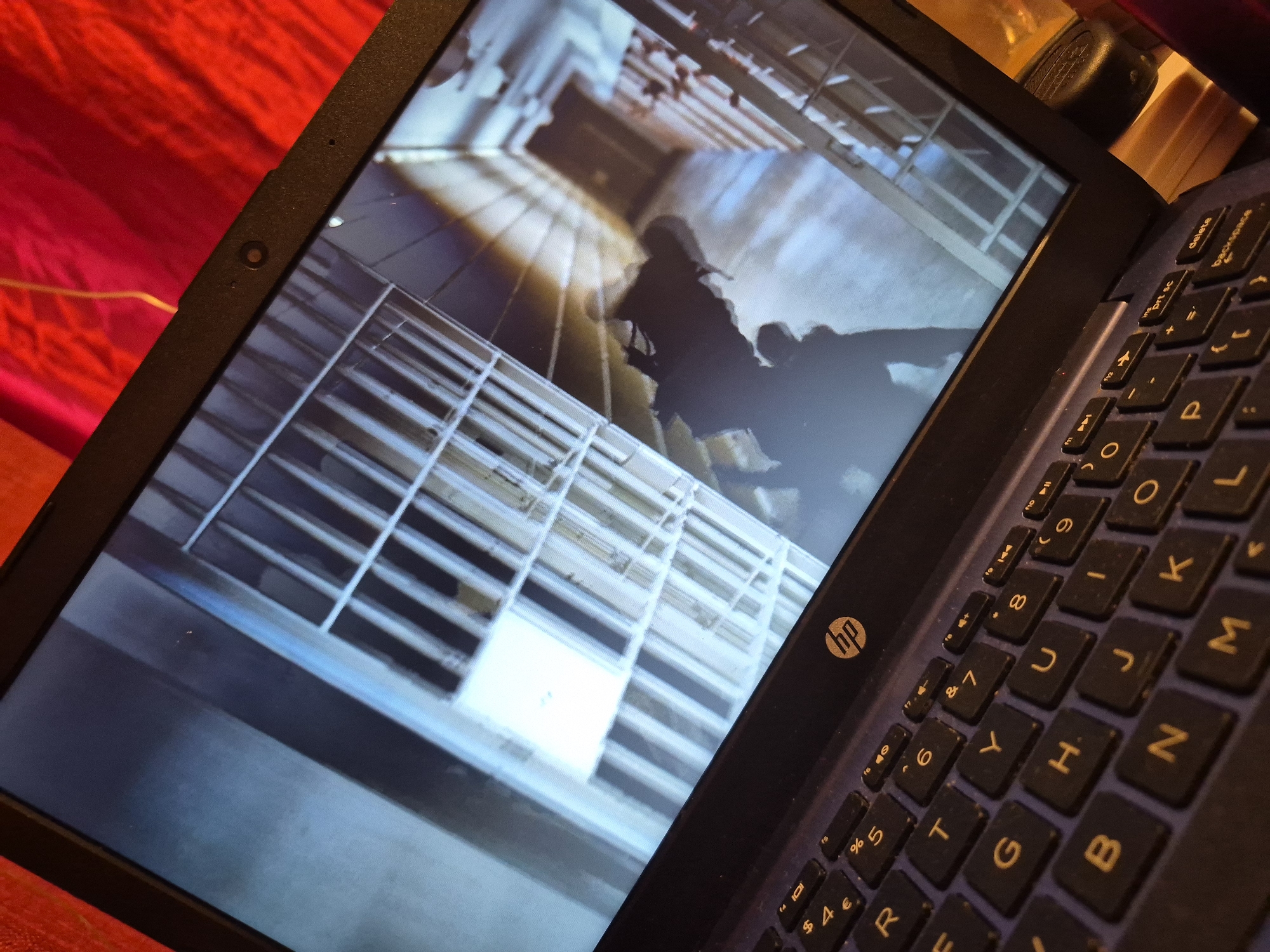 A laptop is open on a dark blue surface, possibly a desk or table, with a bright red fabric draped behind it. The laptop screen displays a scene that appears to be a dimly lit, sterile hallway with metal bars, reminiscent of a prison or secure facility. The perspective is looking down the hallway, with a bright light source at the far end, casting long shadows. A silhouette of a person, or possibly multiple figures, can be seen walking or standing in the hallway. The keyboard of the laptop is visible in the foreground, showing the HP logo and various keys. The surrounding environment is out of focus, but the red fabric suggests an indoor setting. There is no information in the image that indicates the location in the city of Sandwell, United Kingdom.