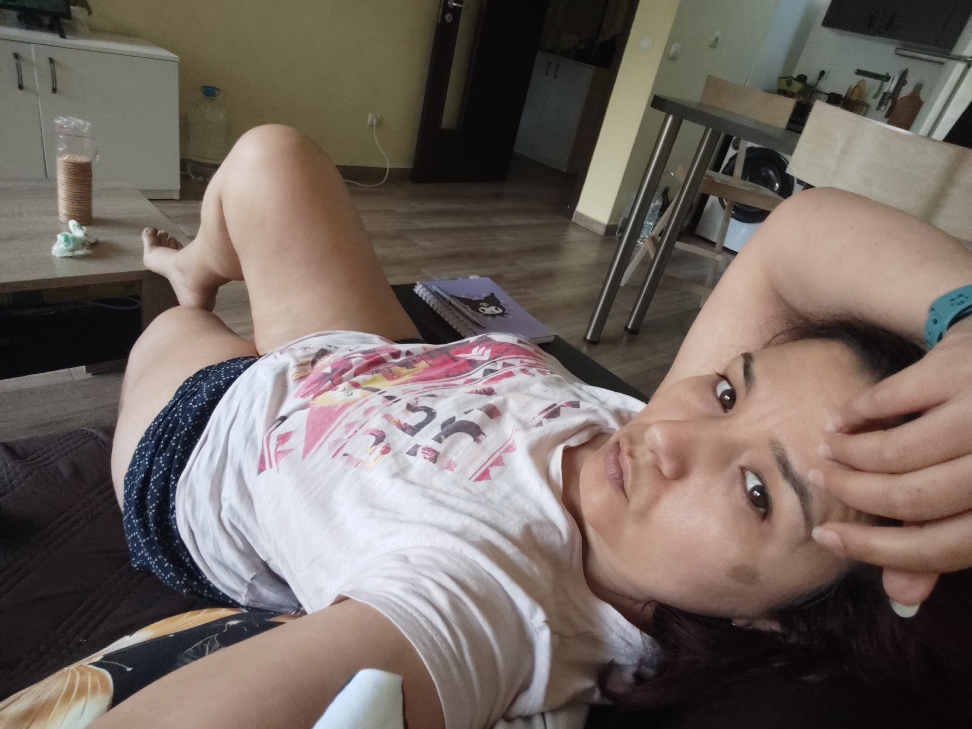 The image shows a woman lying on her side on a dark surface, possibly a sofa or bed. She is wearing a white t-shirt with a colorful, abstract pattern and dark blue shorts with white polka dots. Her left arm is bent and resting behind her head, while her right hand is touching her face. She is looking towards the camera with a neutral expression. Her hair is dark and spread out.
The background appears to be an indoor setting, likely a living room or bedroom. To the left, a wooden coffee table is visible with a plastic container of cookies and some crumpled white paper. Behind the table, a white cabinet with two doors and a bottle of water are present. The floor is made of wood-look laminate. Further in the background, a dark door, a kitchen counter with various items, and part of a dining table with metal legs can be seen. The overall lighting suggests it is daytime. The location context is Plovdiv, Bulgaria.