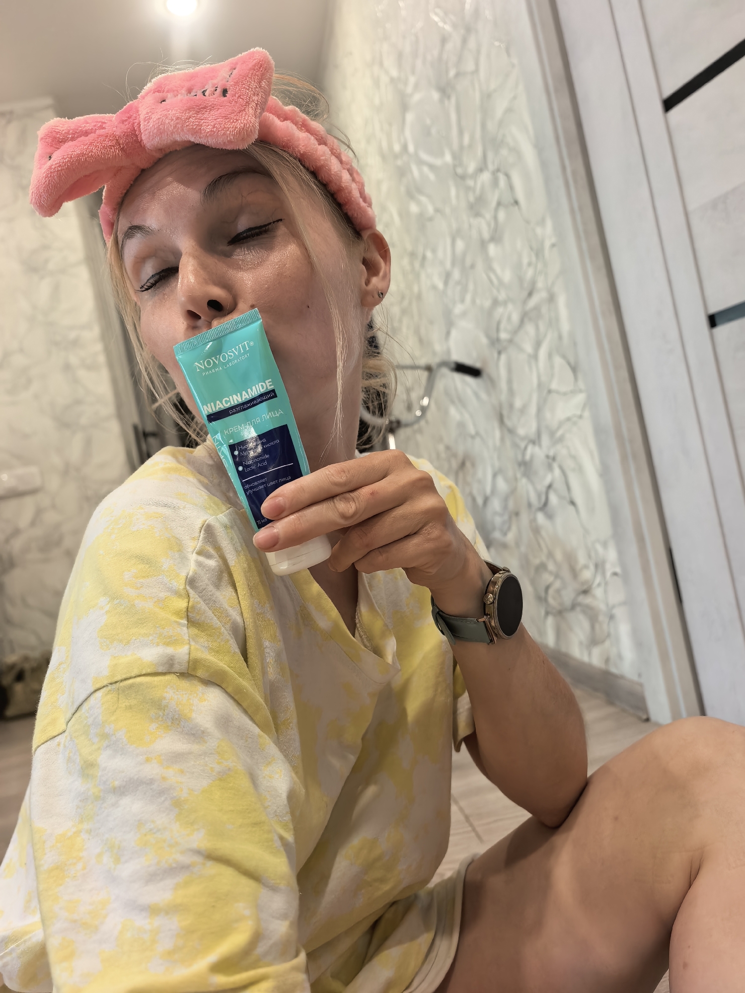 A fair-skinned woman with blonde hair is indoors, sitting on the floor. She's wearing a pink headband and a yellow and white tie-dye t-shirt. The woman has her eyes closed and is holding a blue and white tube of "NOVOSVIT® NIACINAMIDE" face cream close to her face. She is wearing a smart watch on her left wrist. The background includes a patterned wall and a white door with black accents. The location is in Sterlitamak, Russia, as indicated by the Cyrillic text "разглаживающий КРЕМ ДЛЯ ЛИЦА" on the cream tube, which translates to "smoothing face cream."