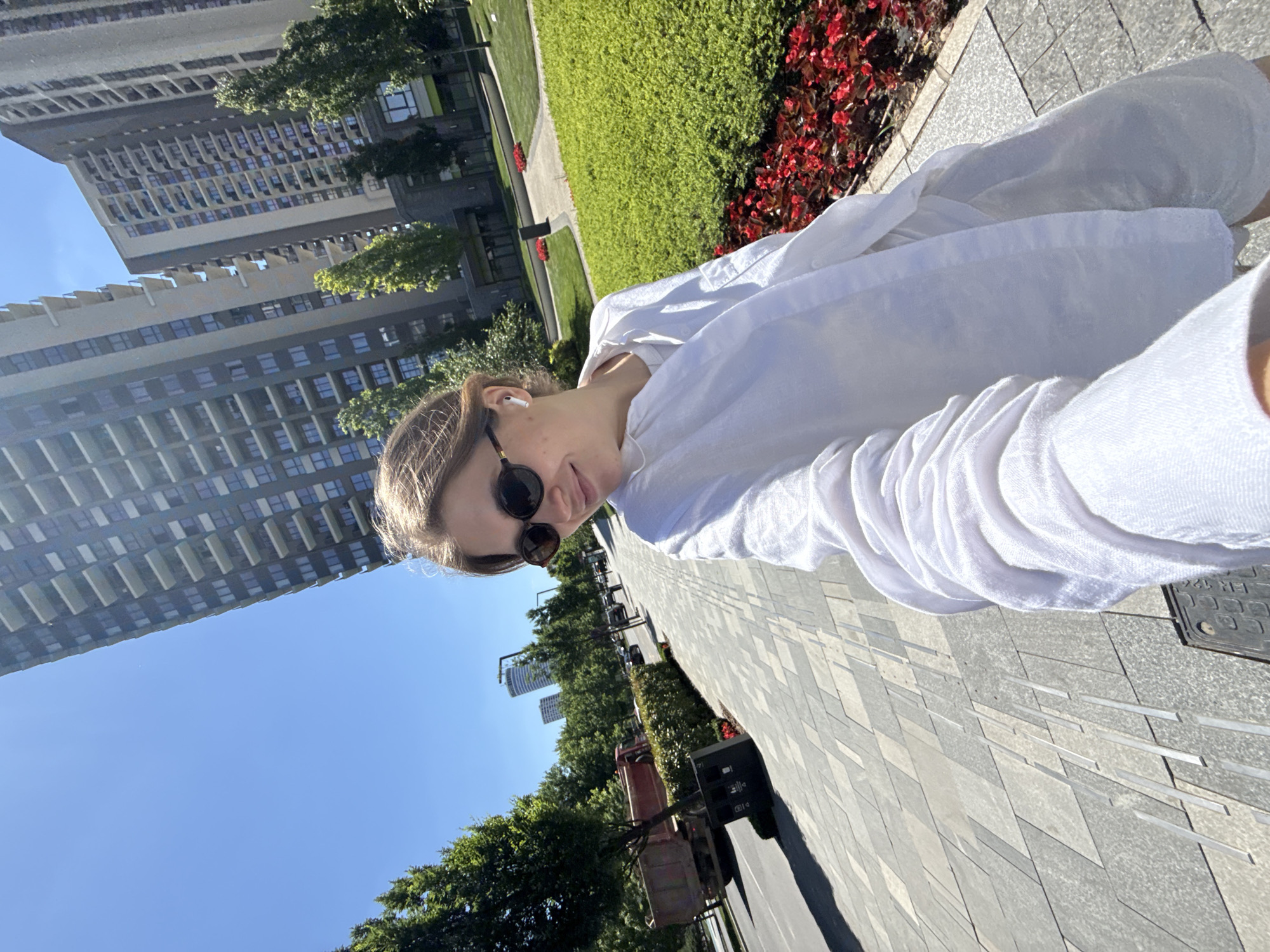 A selfie captures a fair-skinned woman in sunglasses and a white blouse against an urban backdrop. The location appears to be Belgrade, Serbia, given the architecture. In the background, tall, modern buildings rise into a blue sky, interspersed with green trees. A paved walkway stretches towards the horizon, bordered by greenery and a vibrant red flowerbed. A utility vehicle is also visible.