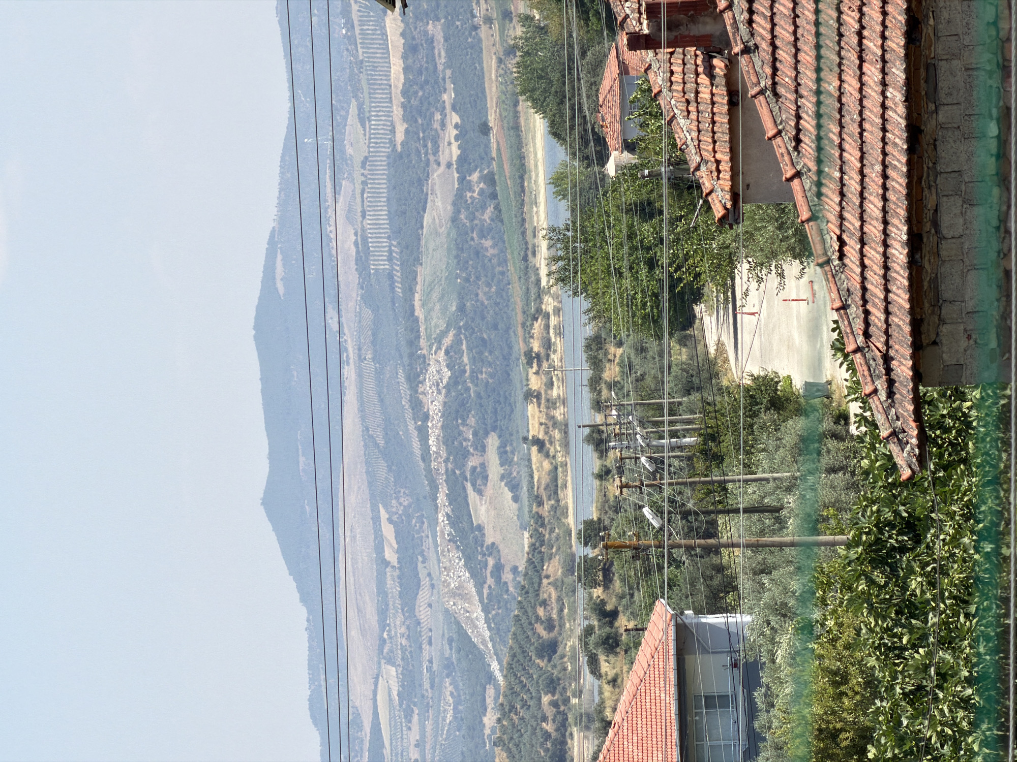 A wide-angle, eye-level, outdoor, daytime, full shot captures a rural landscape under a clear, pale blue sky. In the foreground, the terracotta-tiled roofs of two houses dominate the bottom half of the frame. The house on the left has white-painted walls and a visible window. The house on the right is partially visible, showing a brick chimney and a wall constructed of concrete blocks. Dense green foliage fills the space between and around these structures.
In the mid-ground, a road curves through a small village. Several utility poles with electrical wires stretch across the scene, and a few orange traffic cones are visible on the roadside. Beyond the village, a body of water, possibly a lake or reservoir, is nestled between rolling hills.
The background features a vast, layered landscape of hills and mountains. The slopes are covered in a patchwork of vegetation, including olive groves, indicated by the orderly rows of trees. The distant mountains fade into a hazy, light blue silhouette against the sky. The overall impression is a tranquil rural setting in the vicinity of Köprübaşı, Turkey.