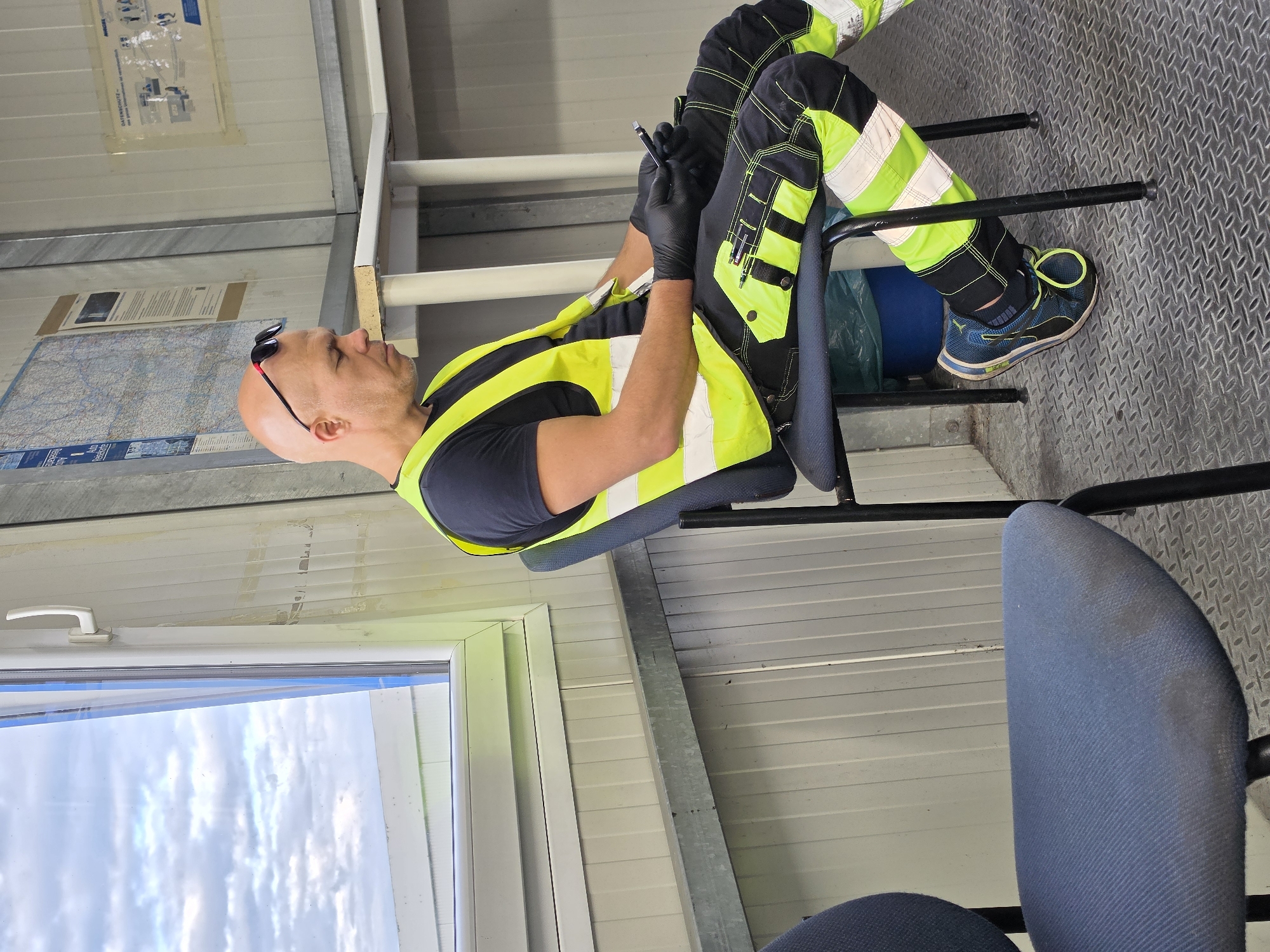 A bald man wearing black gloves, a dark t-shirt, a bright yellow safety vest, and black and neon green work pants is sitting on a dark blue office chair in a room with corrugated metal walls. He is holding a black smartphone in his gloved hands and appears to be looking at it. He has sunglasses perched on his head. The room has a large window with a white frame, showing a cloudy sky and a building in the distance. There is another empty blue office chair in the foreground. On the wall behind the man, there are several papers and maps. A blue trash can is visible under the chair. The floor is a textured metal diamond plate pattern. The man is likely on a break at a worksite. Based on the maps and signage, this location appears to be Flörsheim, Germany.